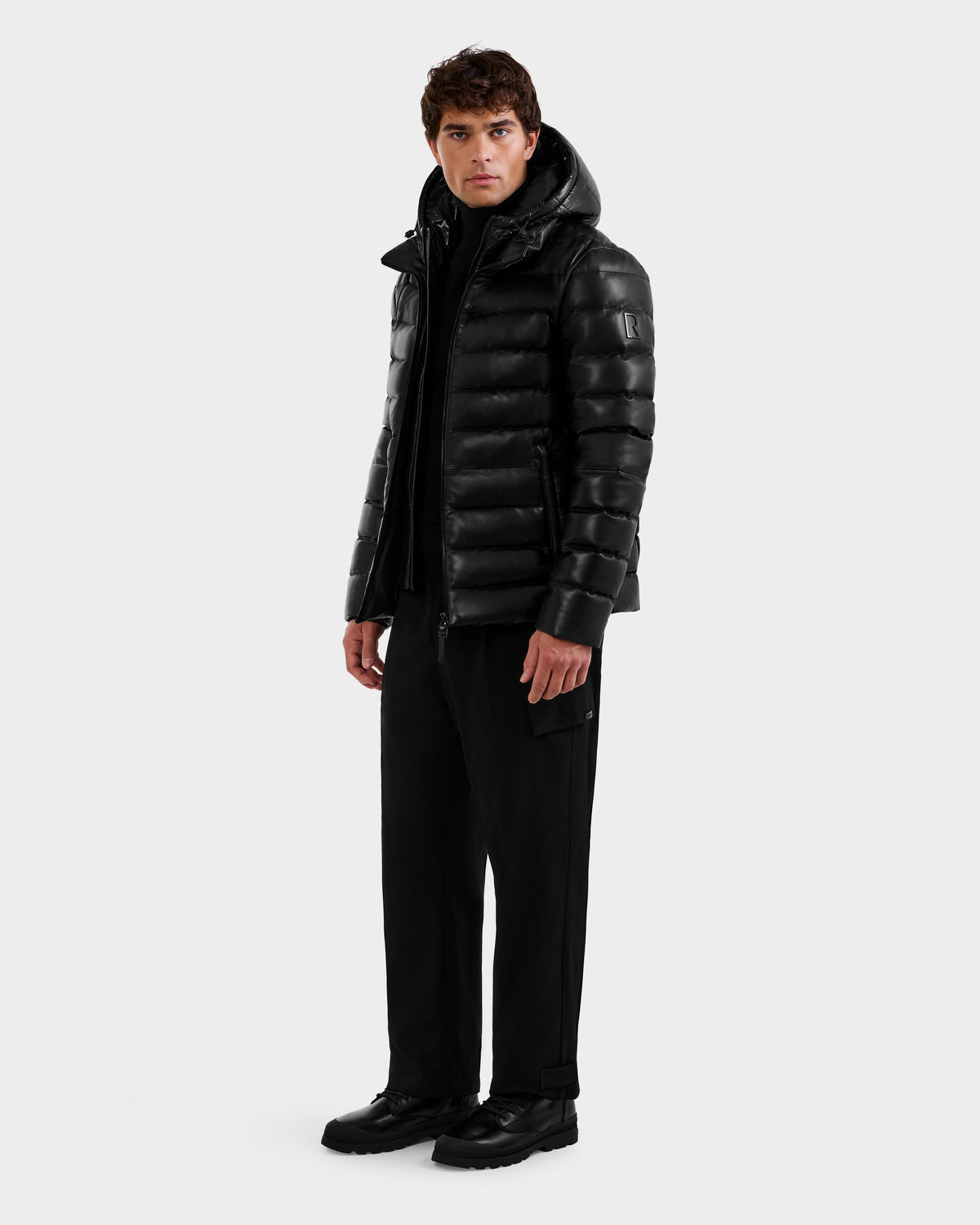 A man stands facing forward against a light gray background, wearing the Rudsak DRAVENX - 6125686 Black leather puffer jacket with a hood, paired with black pants and black shoes.