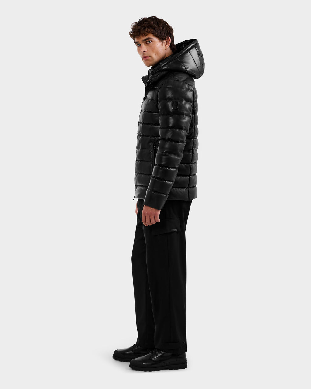 A young man wearing the Rudsak DRAVENX - 6125686 Black leather puffer jacket with a hood, black pants, and black shoes stands angled slightly to the side against a plain light background.