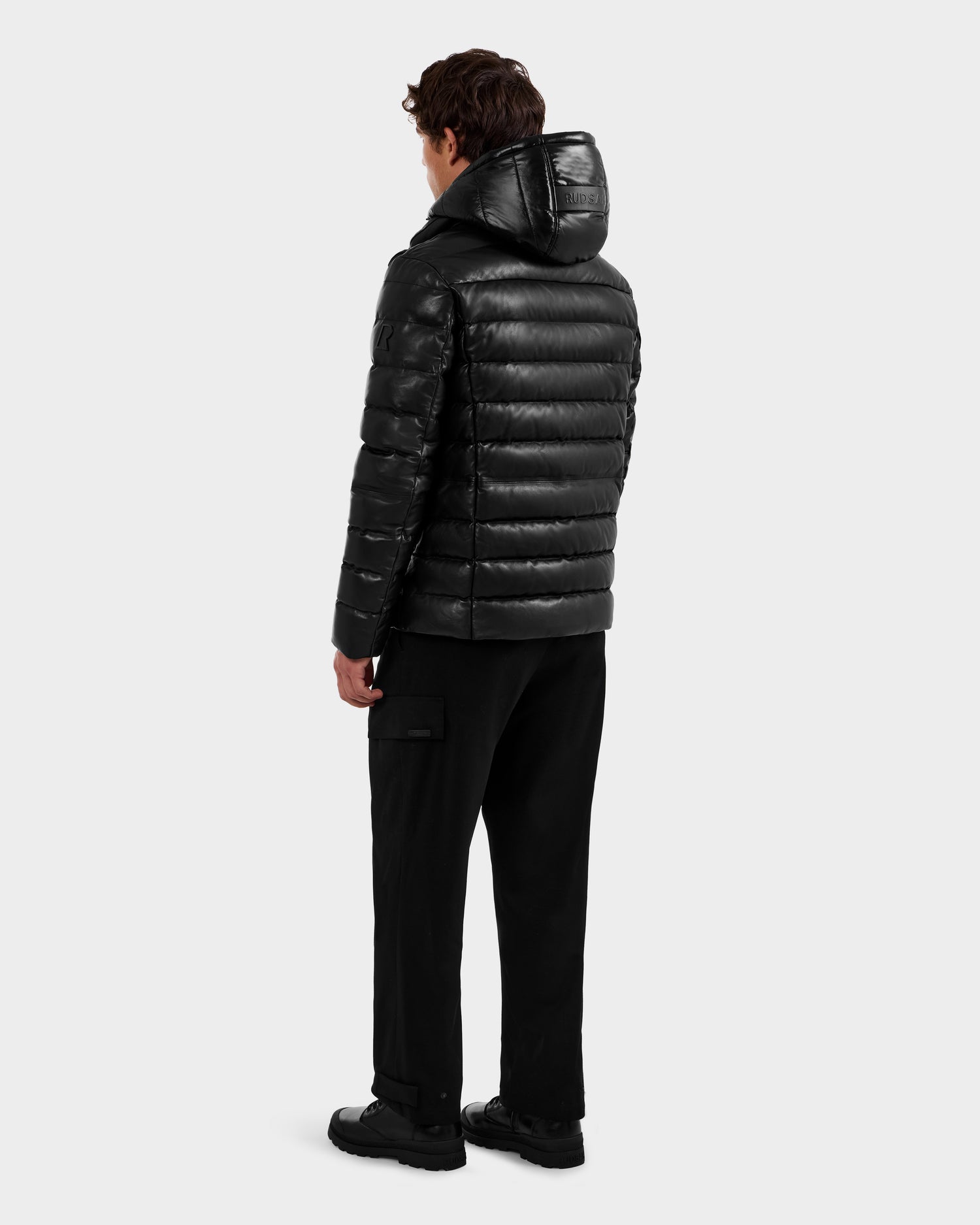 A person stands facing away, wearing a shiny black Rudsak DRAVENX - 6125686 men’s leather down jacket with a hood, paired with black pants and black shoes, against a plain light gray background.