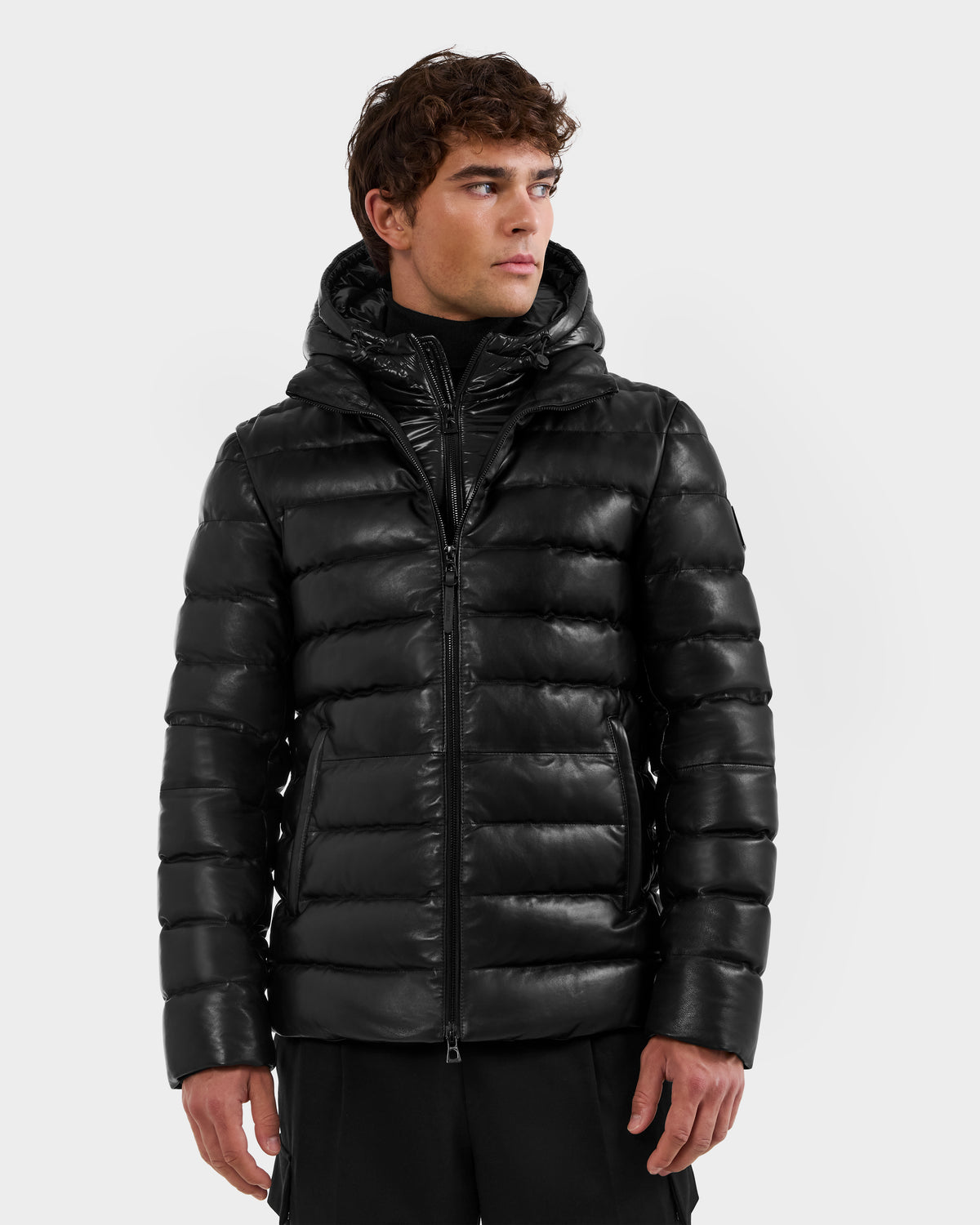 A man in a black Rudsak DRAVENX - 6125686 leather puffer jacket with a hood stands against a plain light background, looking left with a neutral expression.