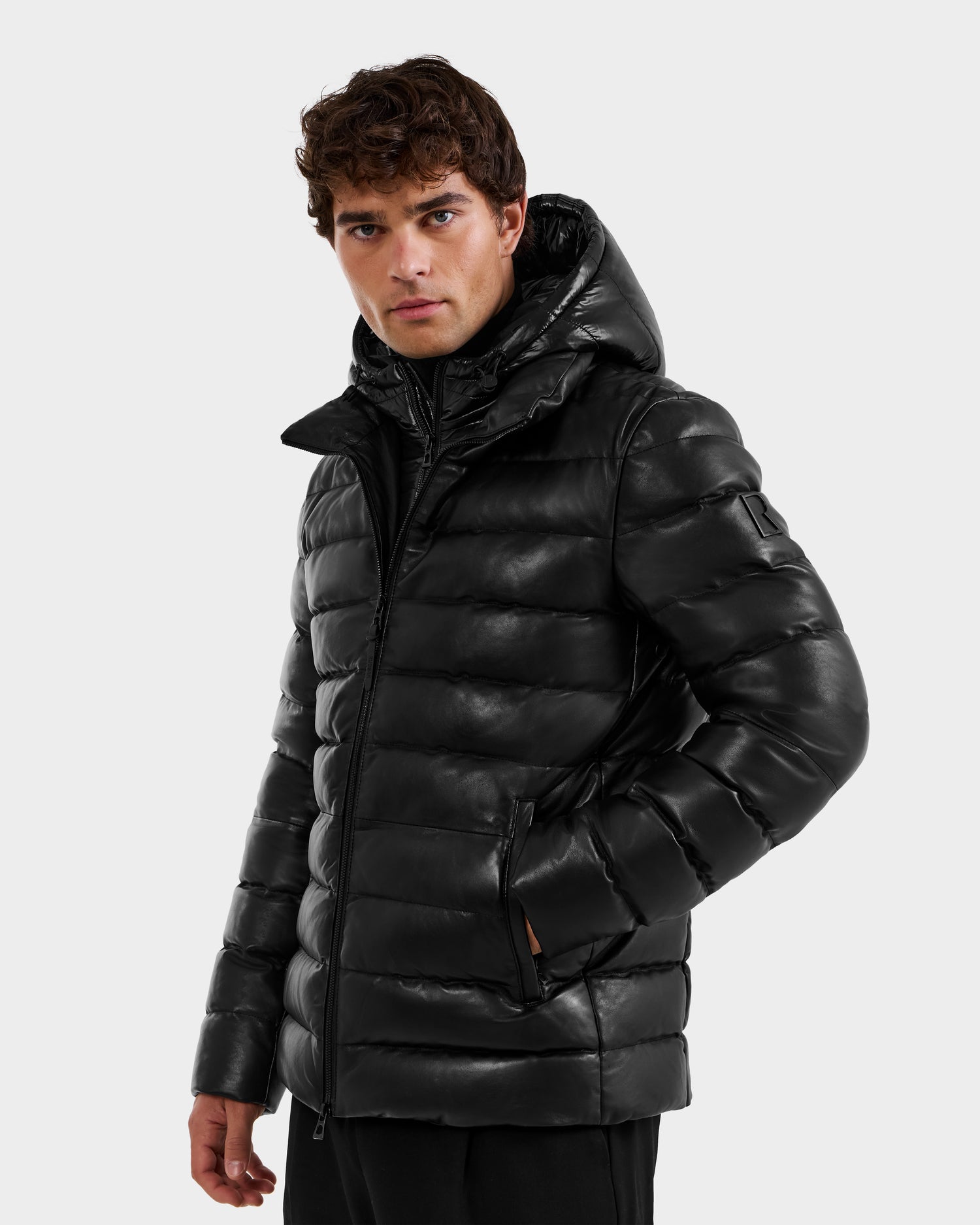 A man with short curly hair, looking slightly to the side, wears the Rudsak DRAVENX - 6125686 Black shiny leather puffer jacket with a hood, standing against a plain light gray background.