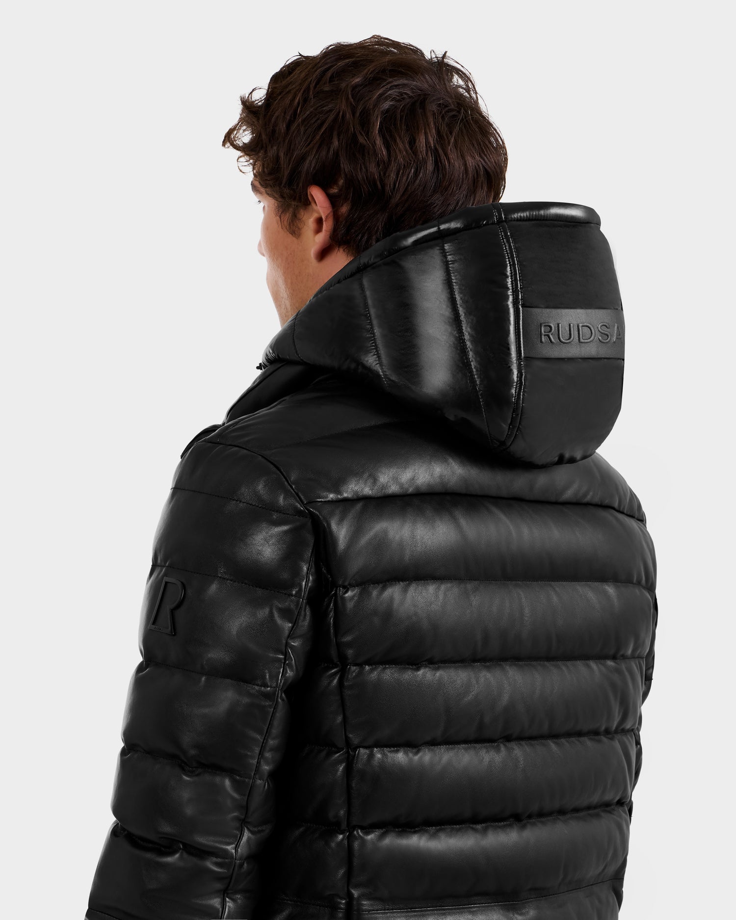 A person with short brown hair is seen from behind wearing the shiny black Rudsak DRAVENX - 6125686 leather puffer jacket. "RUDSAK" is embossed on the large hood, and the background is plain.