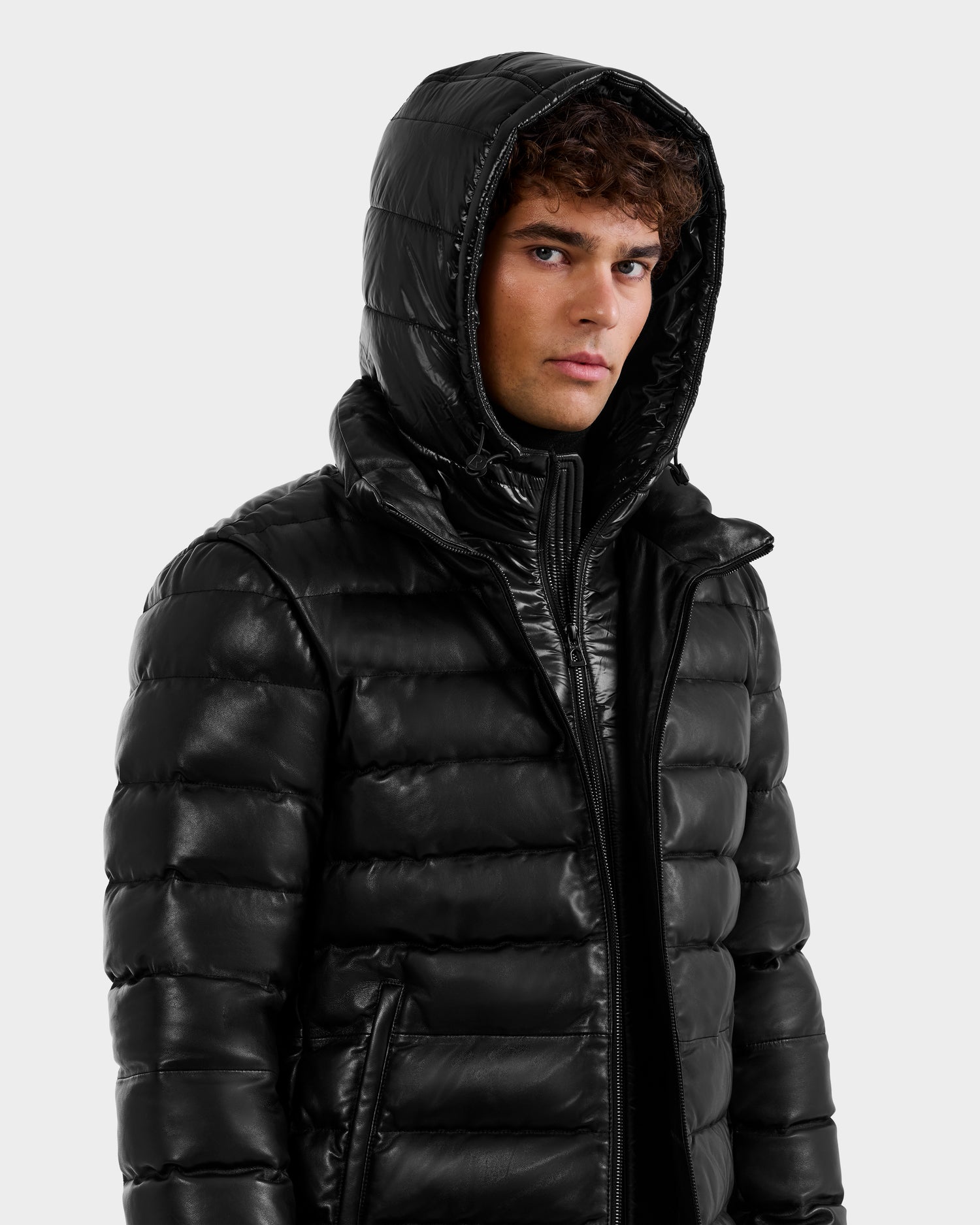 A young man with curly brown hair wears the Rudsak DRAVENX - 6125686 Black leather puffer jacket with a hood, standing slightly to the side against a plain light background while looking at the camera.