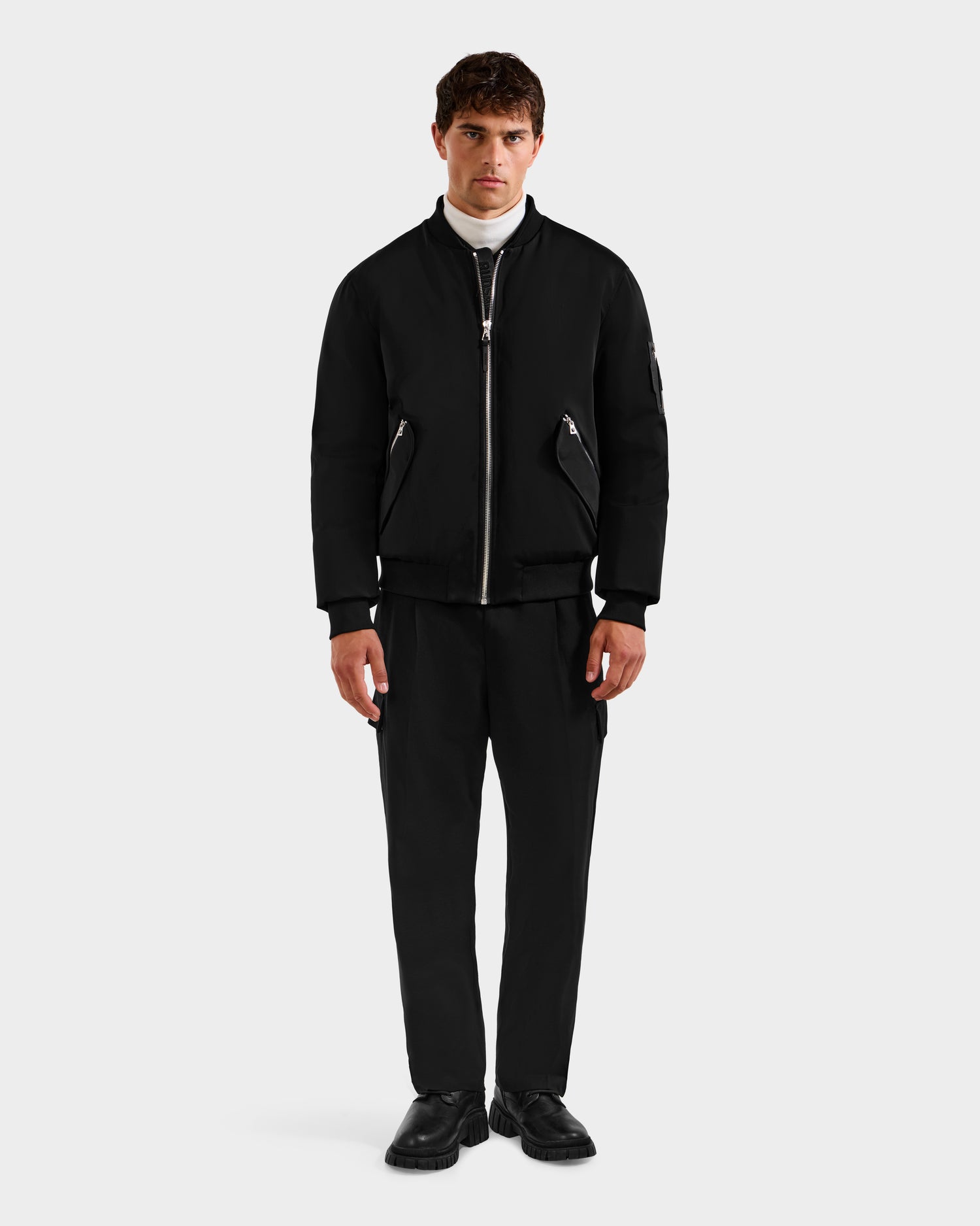 A man faces forward wearing the Rudsak DYLAN - 6125593 Black aviator bomber jacket with a removable faux fur collar, styled with black pants, black shoes, and a white turtleneck against a plain light gray background.