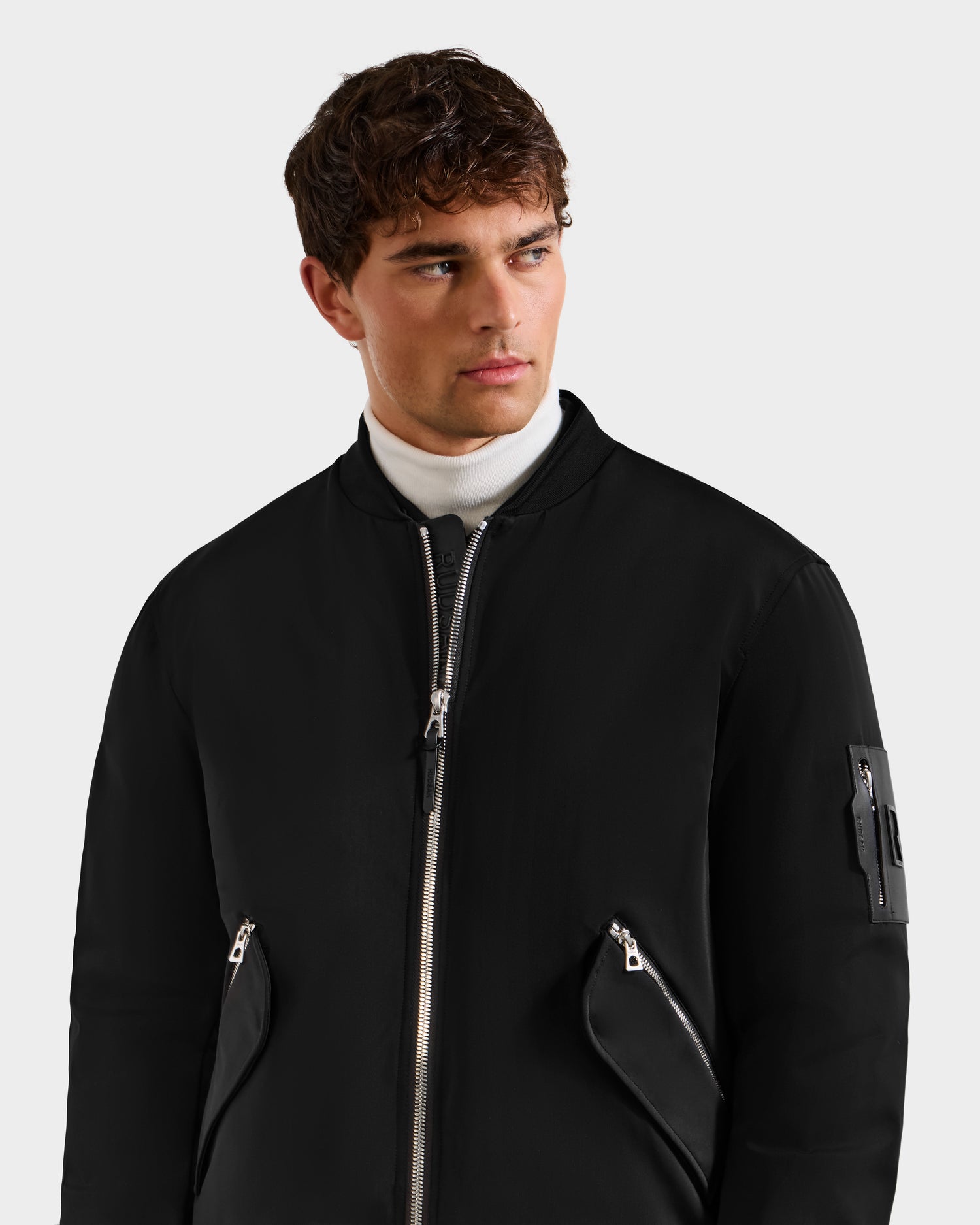 A man in the Rudsak DYLAN - 6125593 Black men’s aviator bomber jacket with zippered pockets and a removable faux fur collar, layered over a white turtleneck, looks left against a plain light background.
