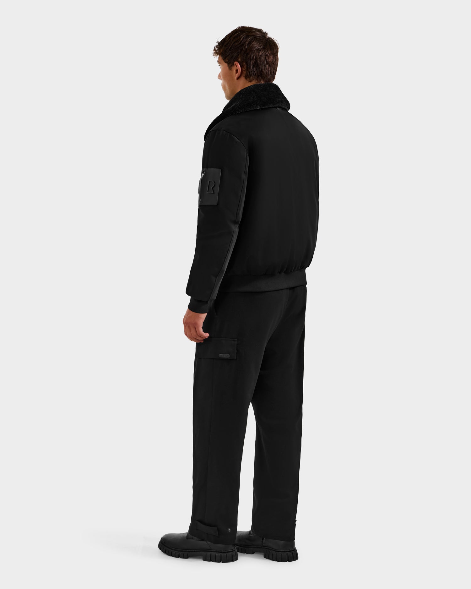 A man faces away, wearing the Rudsak DYLAN - 6125593 Black men’s aviator bomber jacket with a removable faux fur collar, paired with black cargo pants and shoes against a plain light background.