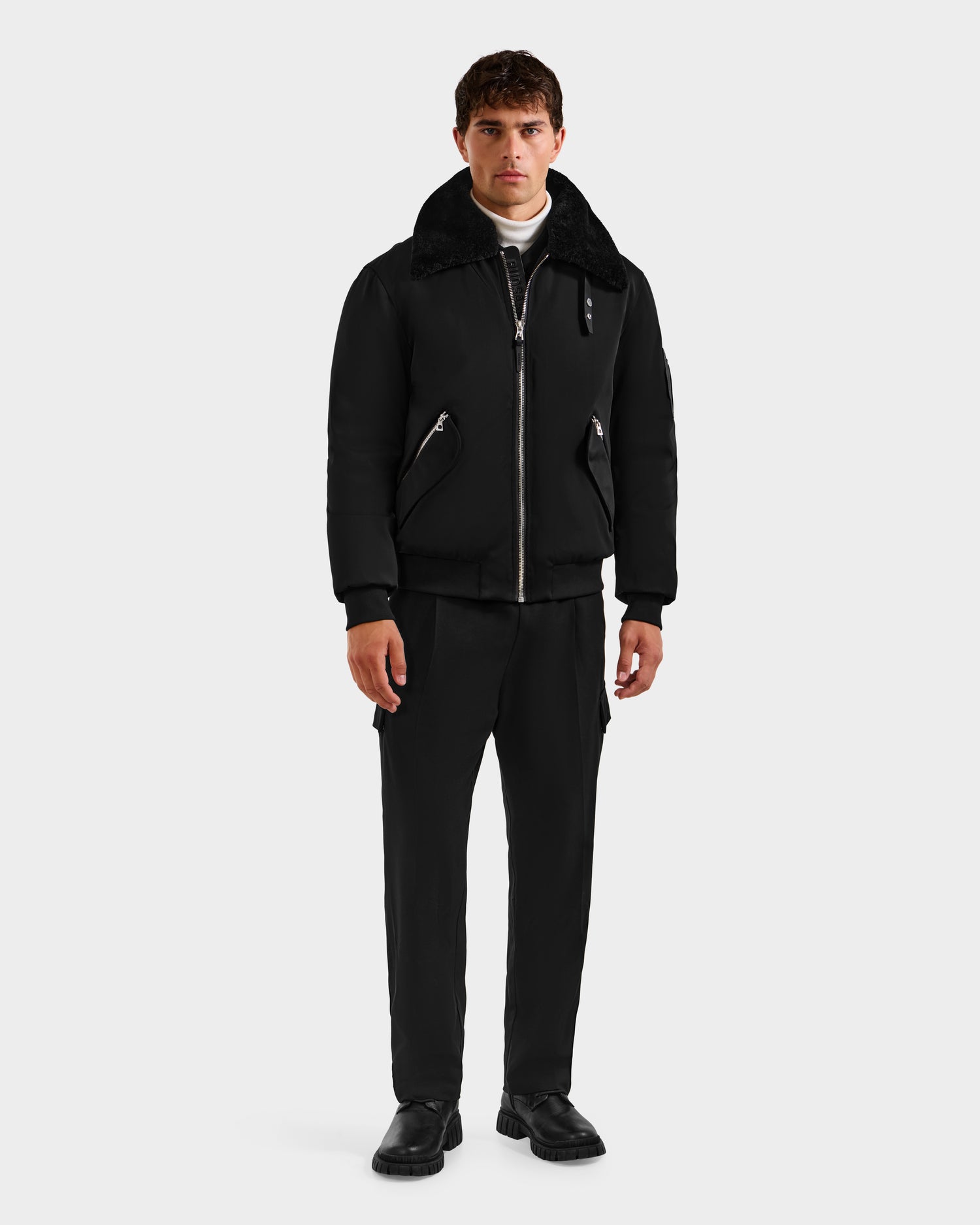 A man faces forward, wearing the Rudsak DYLAN - 6125593 Black men’s aviator bomber jacket with a removable faux fur collar, paired with black pants, black shoes, and a white turtleneck against a plain light background.