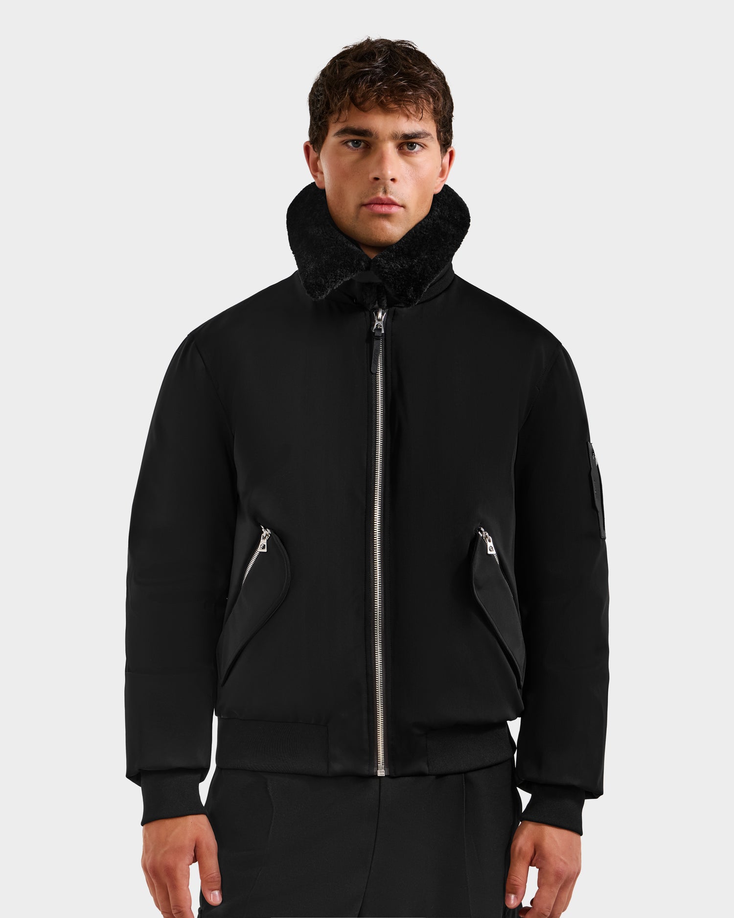 A person in the Rudsak DYLAN - 6125593 Black men’s aviator bomber jacket with a removable faux fur collar and zippered pockets stands against a light gray background, looking directly at the camera.