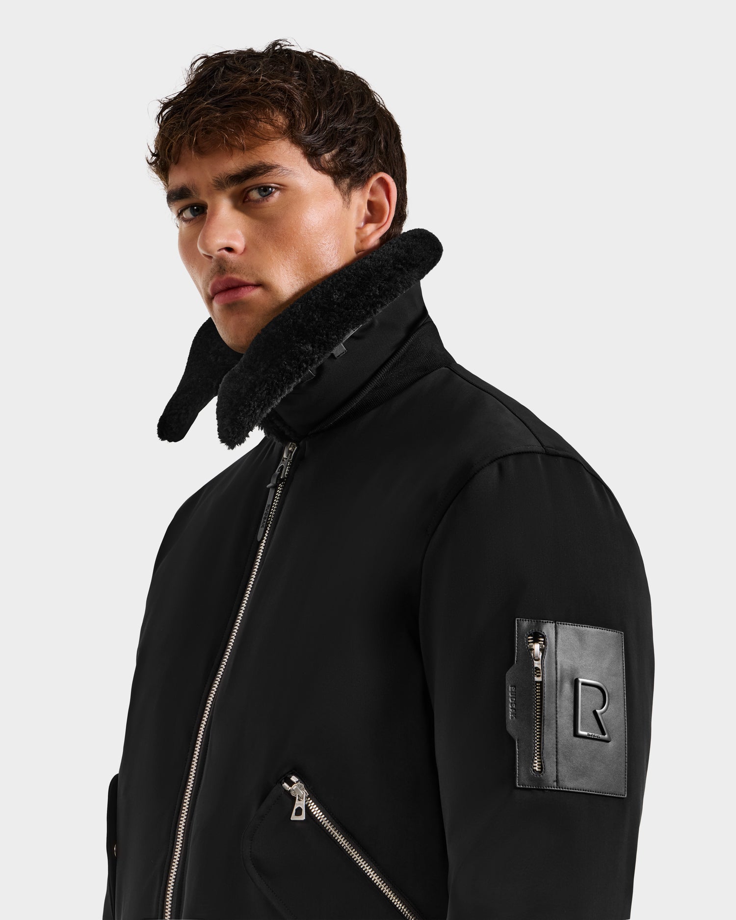 A young man with short brown hair wears the DYLAN - 6125593 Black aviator bomber jacket by Rudsak, featuring a high shearling collar, zippered pockets, and an embossed R leather patch on the sleeve. He looks seriously toward the camera.
