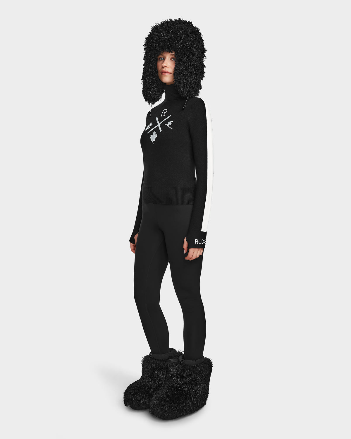 Wearing the ELIZABETH - 8524623 Black by Rudsak, a person models a chic winter look with the large fluffy hat, cozy sweater, black leggings, and oversized furry slippers against a white background.