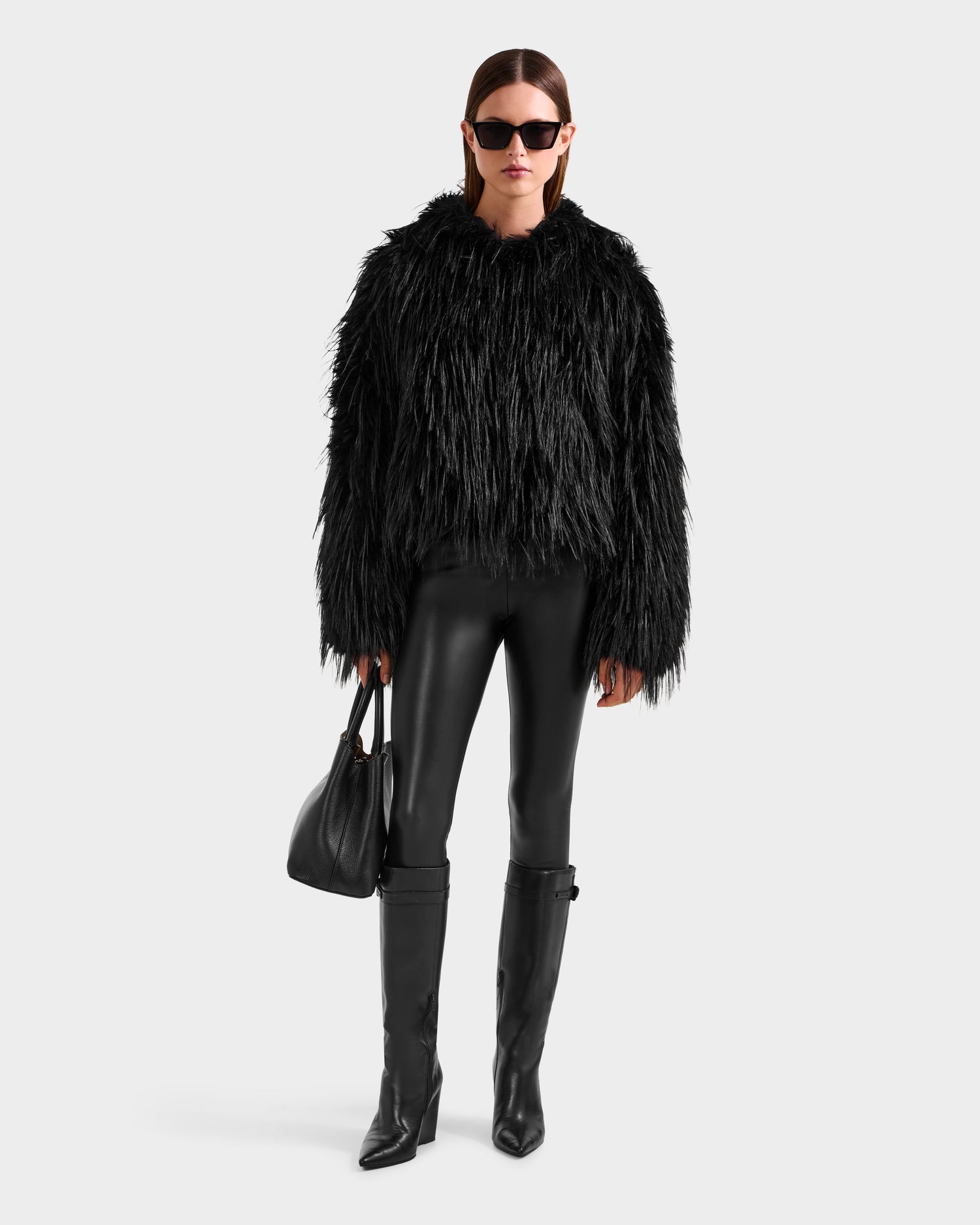 A woman stands against a plain light background, wearing Rudsak's ELLISON - 8525678 Black faux fur hoodie jacket, black leather leggings, knee-high boots, sunglasses, and holding a black handbag.