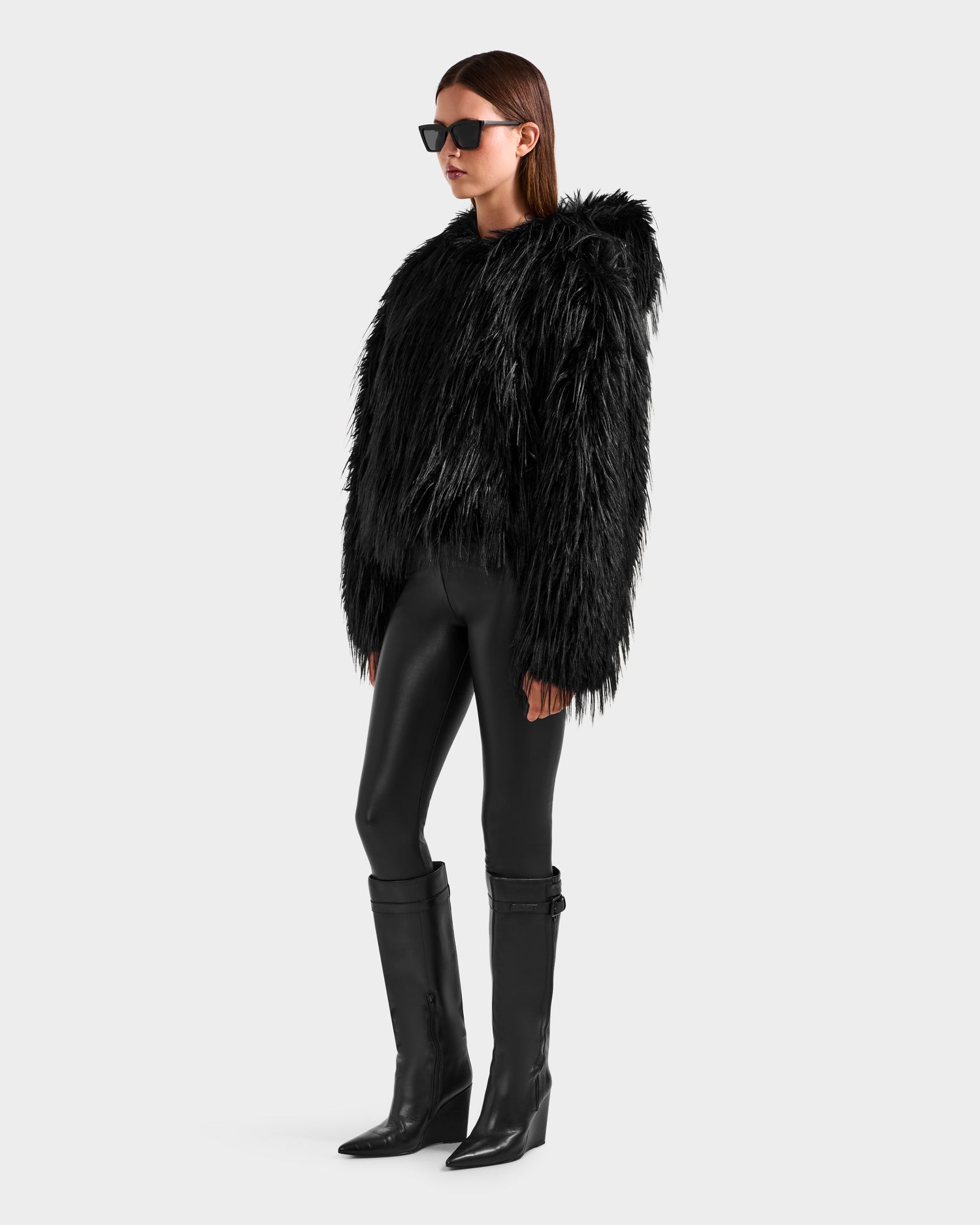 A person models the Rudsak ELLISON - 8525678 Black shaggy faux fur hoodie with an oversized hood, styled with black leggings, tall boots, and sunglasses against a light gray background for an après-ski look.