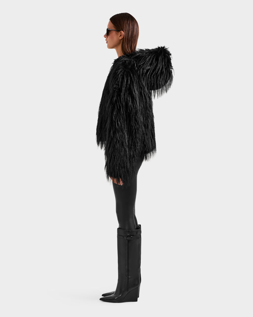 A person in profile stands against a light background, wearing the ELLISON - 8525678 Black textured faux fur hoodie by Rudsak with an oversized hood, paired with black leggings, tall wedge boots, and sunglasses.