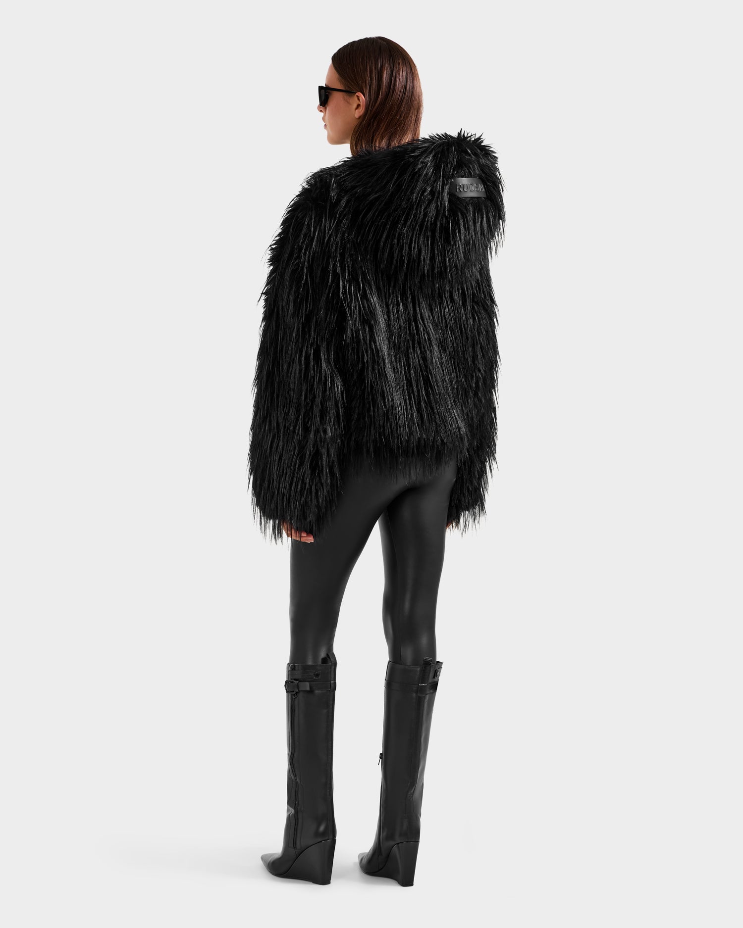 Someone wears the Rudsak ELLISON - 8525678 Black faux fur hoodie with oversized hood, shiny leggings, tall boots, and sunglasses, standing against a light background for a chic après-ski look.