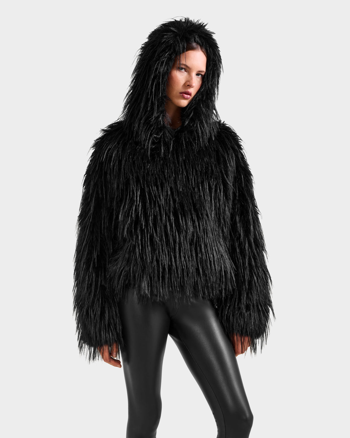 A person in the Rudsak ELLISON - 8525678 Black oversized faux fur hoodie and shiny black leggings stands against a plain light gray background.