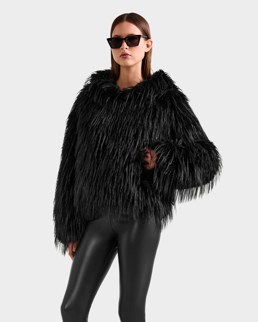 A woman models the Rudsak ELLISON - 8525678 Black faux fur hoodie with an oversized hood, paired with shiny black leggings and black sunglasses, standing against a plain light gray background.