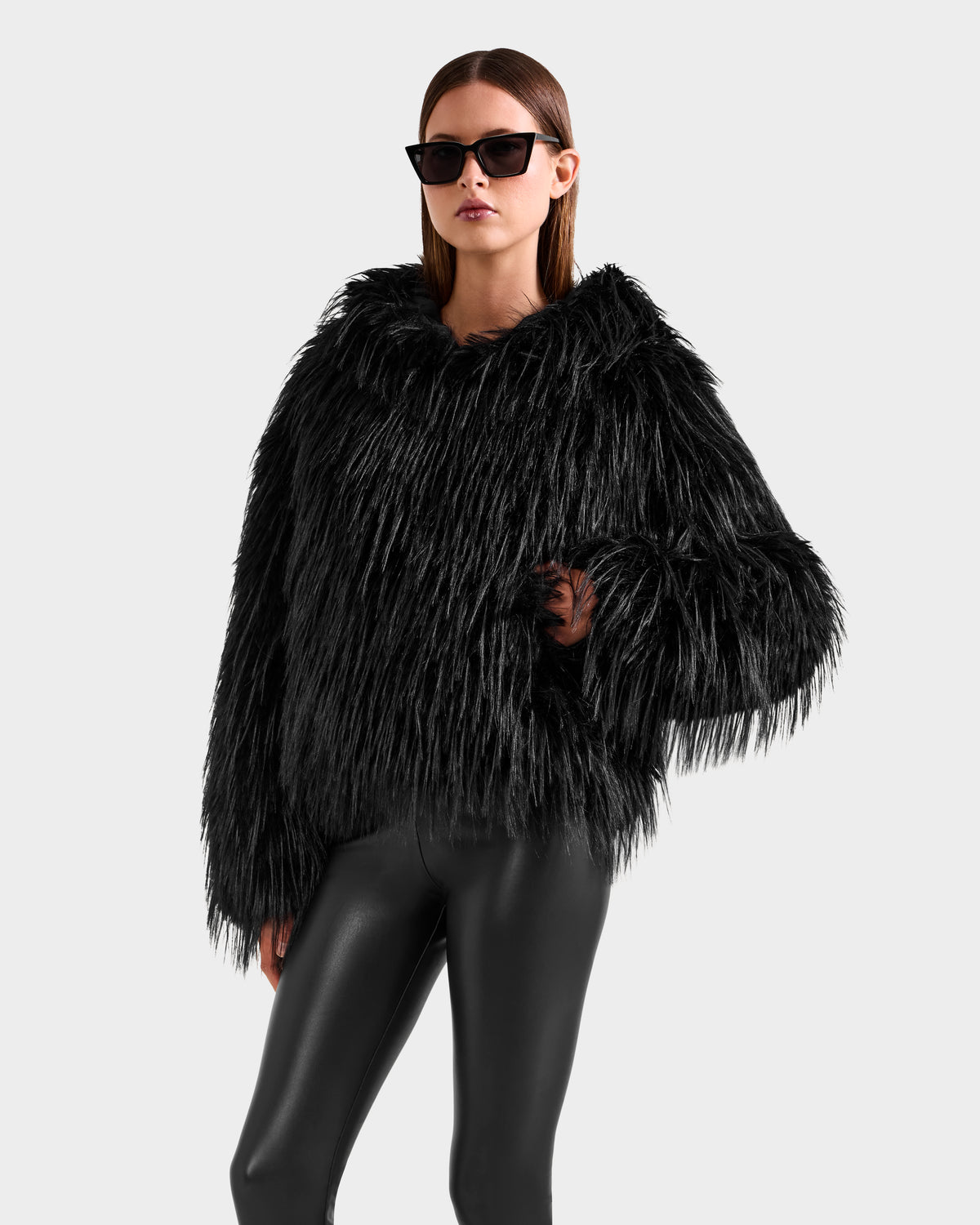 A woman models the Rudsak ELLISON - 8525678 Black faux fur hoodie with an oversized hood, paired with shiny black leggings and black sunglasses, standing against a plain light gray background.