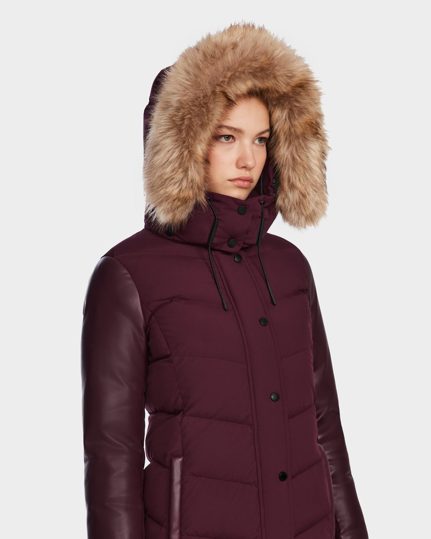 A woman wearing the Rudsak ELSA - L123535 Wine puffer with faux fur-lined hood and leather sleeves stands against a white background, looking forward with a neutral expression.