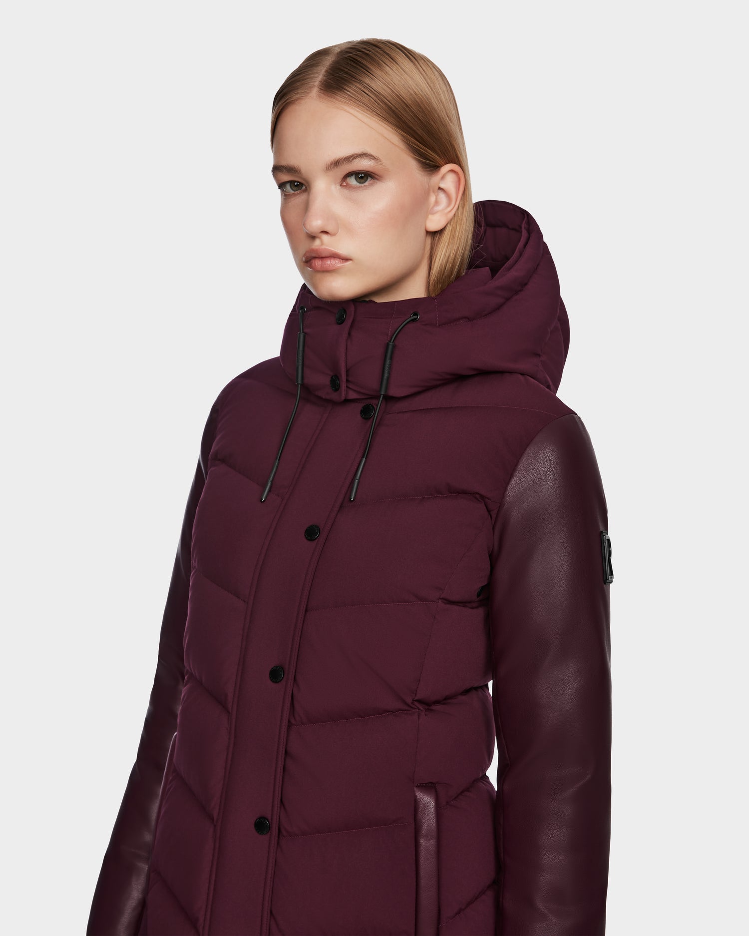 A young woman with straight blonde hair wears the Rudsak ELSA - L123535 Wine quilted down puffer jacket, featuring dark leather sleeves, standing against a plain light background and looking slightly to the side with a neutral expression.