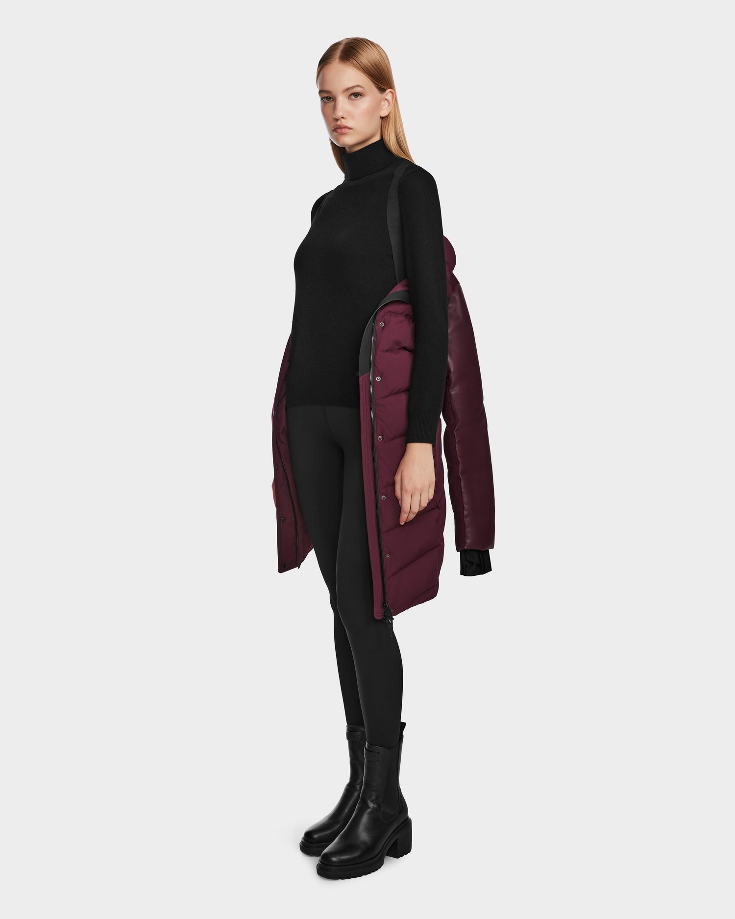 A woman stands against a plain white background with the Rudsak ELSA - L123535 Wine RDS-certified down puffer coat draped over her shoulders, styled with a black turtleneck, leggings, and boots.