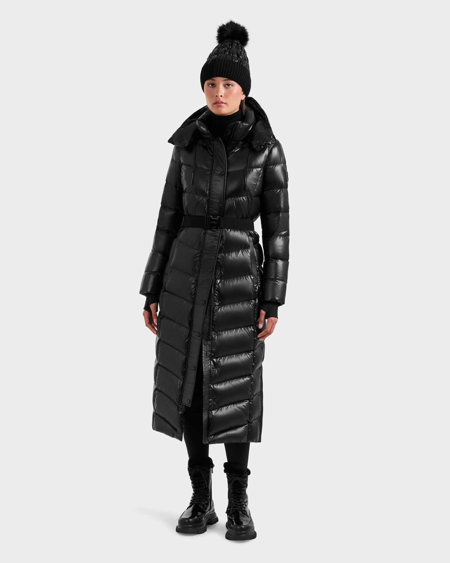 A person wears the Rudsak ELSIE S - L124536 Black women's maxi down puffer coat, black pants, boots, and a pom-pom beanie against a plain light gray background. The stylish coat features responsibly sourced RDS-certified down.