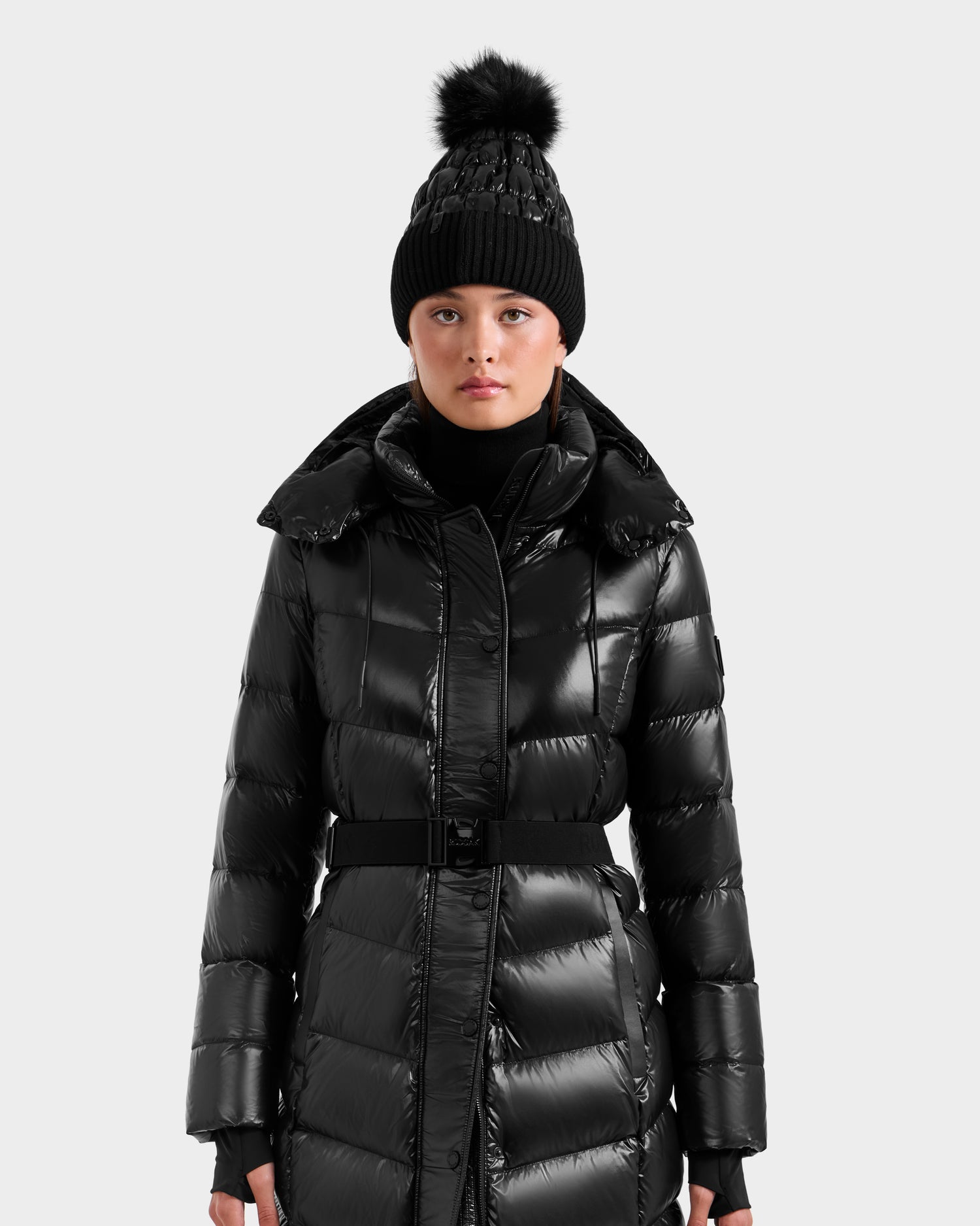 A woman wears the Rudsak ELSIE S - L124536 Black maxi down puffer with a belt and a black beanie topped with a pom-pom, standing against a plain light gray background.
