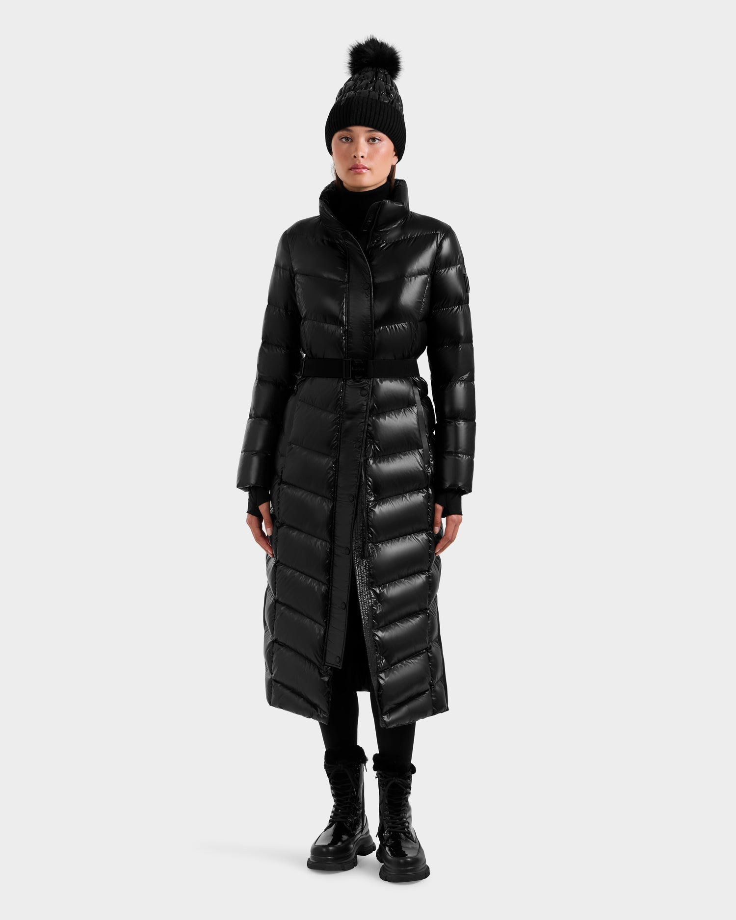 A person in the Rudsak ELSIE S - L124536 Black women’s maxi down puffer, black pants, lace-up boots, and a black pom-pom beanie stands against a plain light gray background.