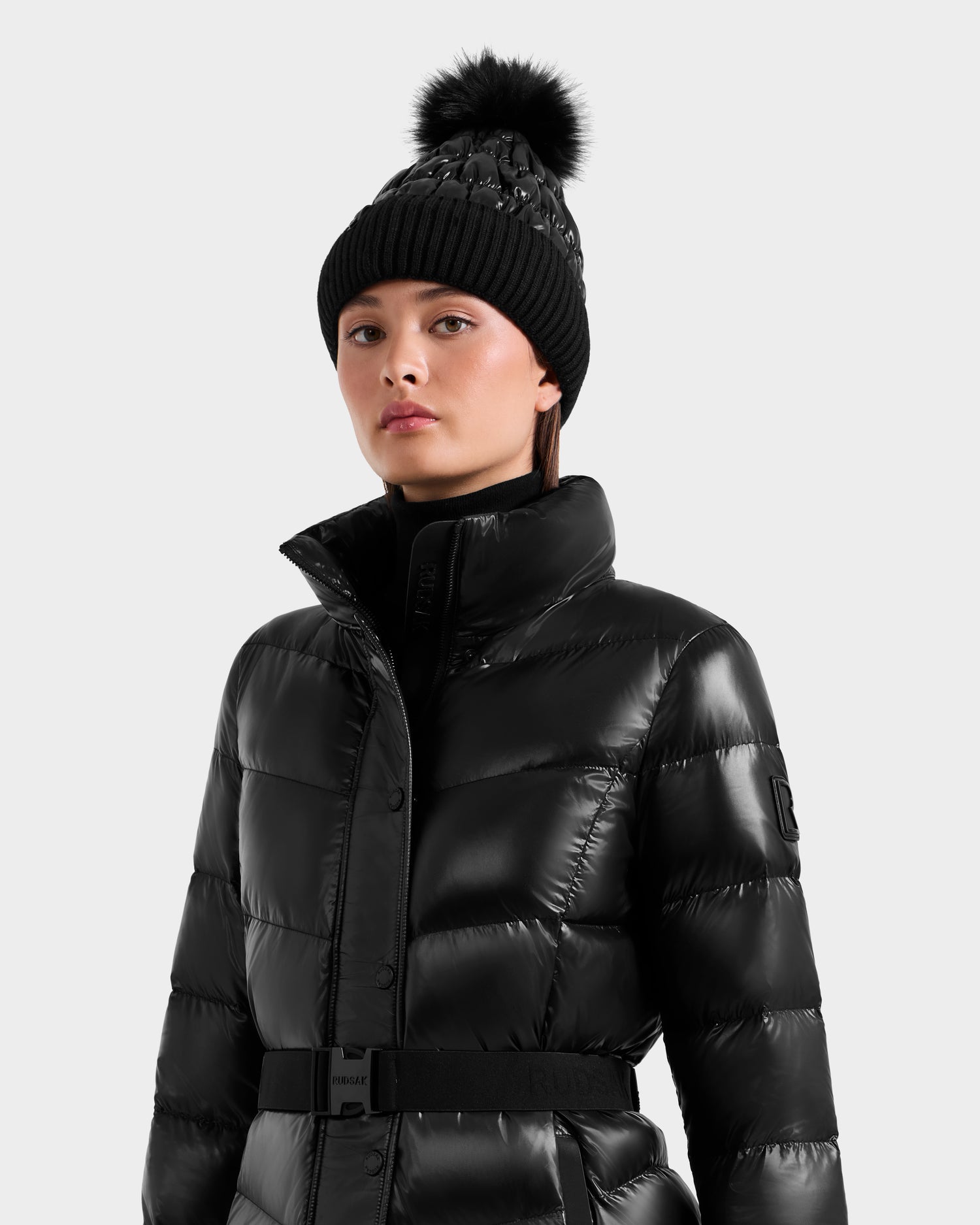 A woman models the Rudsak ELSIE S - L124536 Black puffer jacket with a high collar and a black pom-pom beanie, standing against a light gray background.