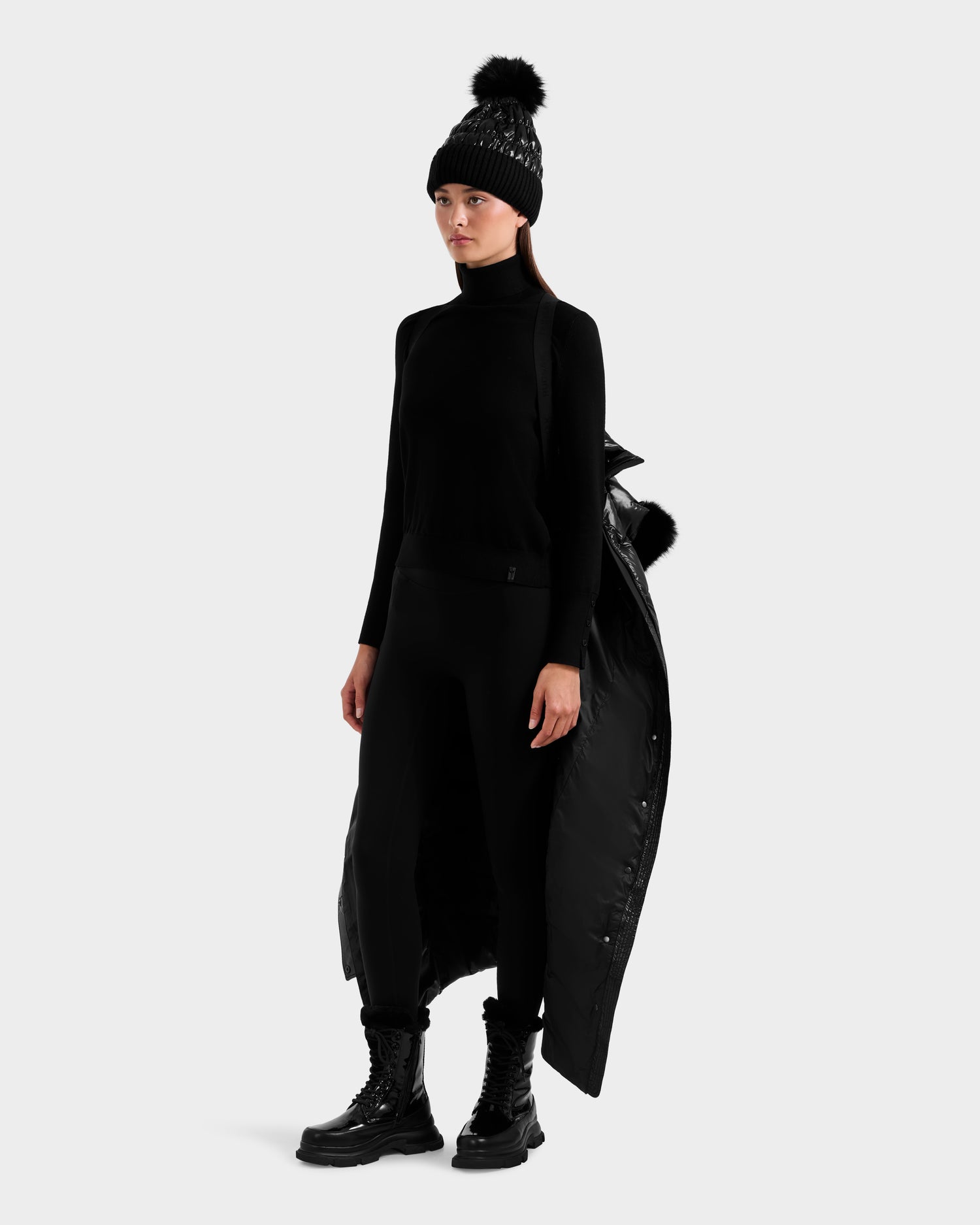 A woman wears an all-black look featuring the Rudsak ELSIE S - L124536 Black maxi down puffer coat with a back fur accent, paired with a knit pom-pom beanie, turtleneck sweater, leggings, and lace-up combat boots.