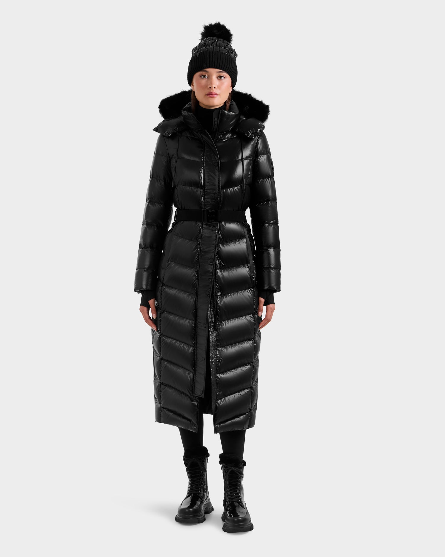 A woman wears the Rudsak ELSIE S - L124536 Black maxi down puffer coat with hood, paired with a black pom-pom beanie, black pants, and boots, standing against a plain white background.