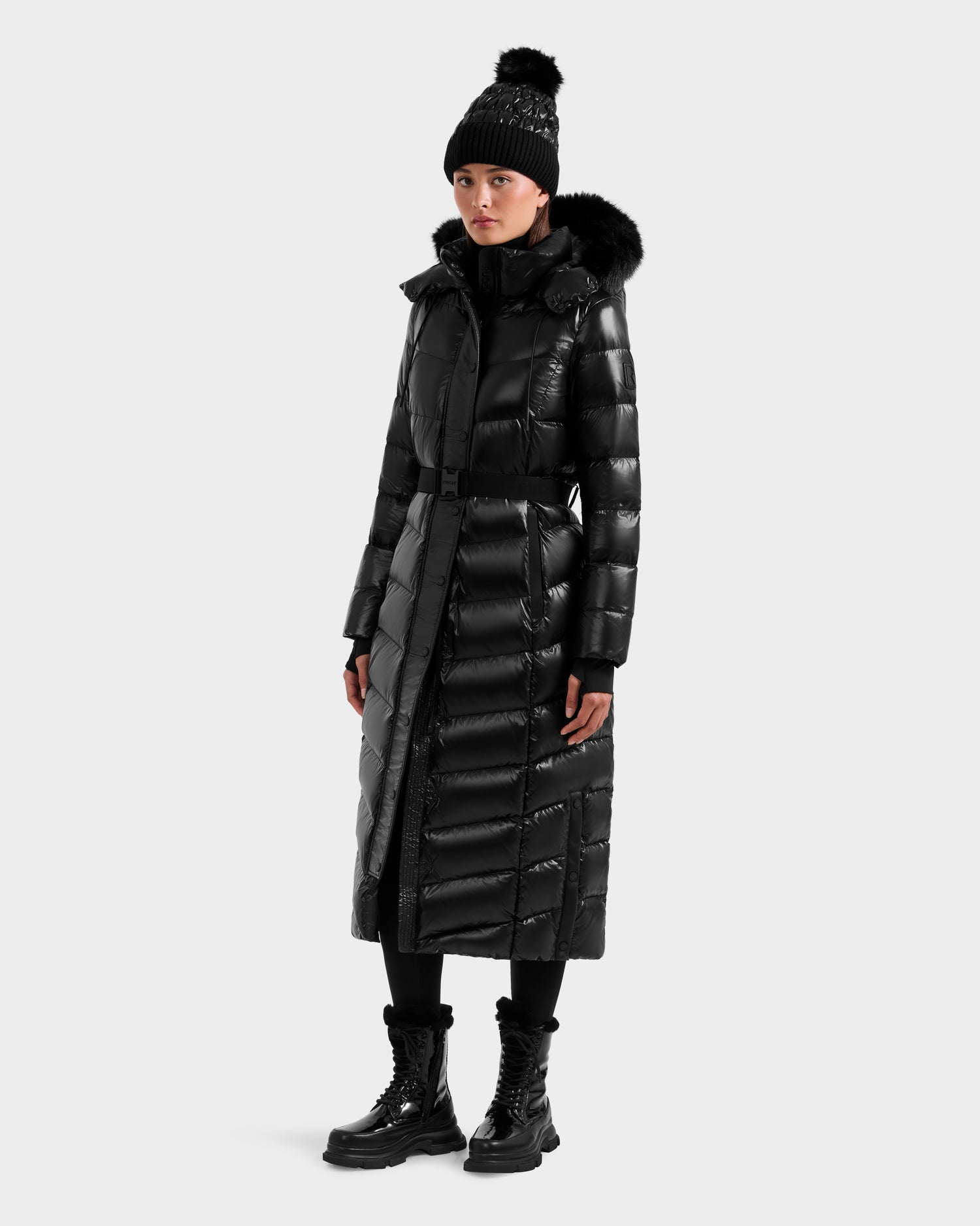 A person wearing the Rudsak ELSIE S - L124536 Black maxi down puffer with a fur-trimmed hood, a black knit hat with pom-pom, black leggings, and lace-up boots stands against a plain light background.
