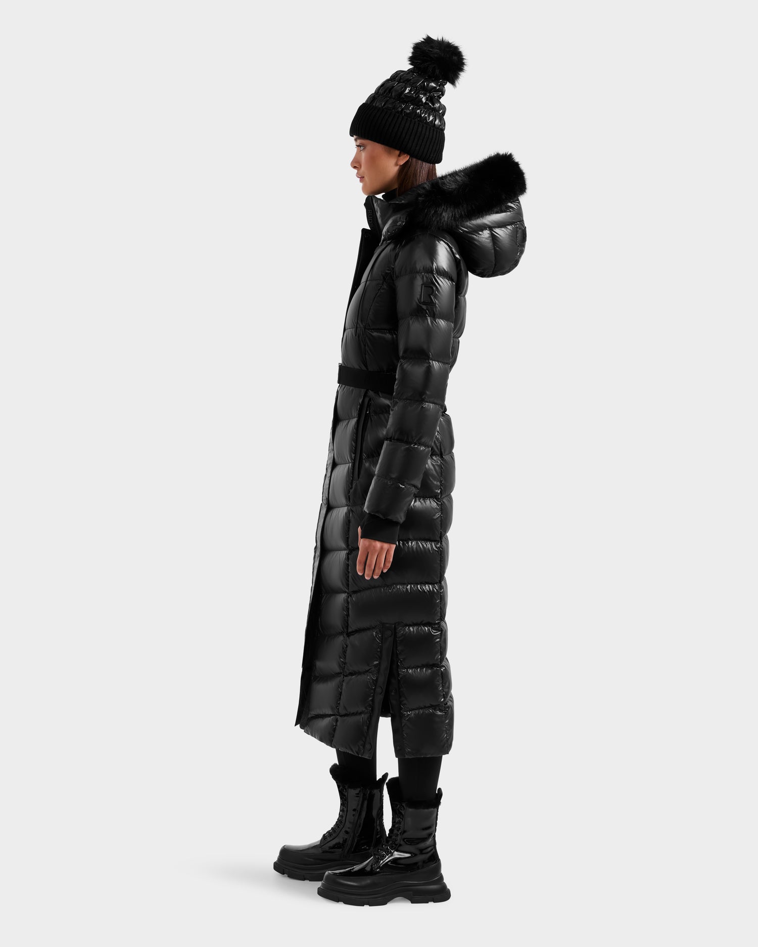 Against a plain light background, a person in profile wears Rudsak's ELSIE S - L124536 Black women’s maxi down puffer winter coat with a fur-trimmed hood, paired with a black pom-pom beanie and black lace-up winter boots.