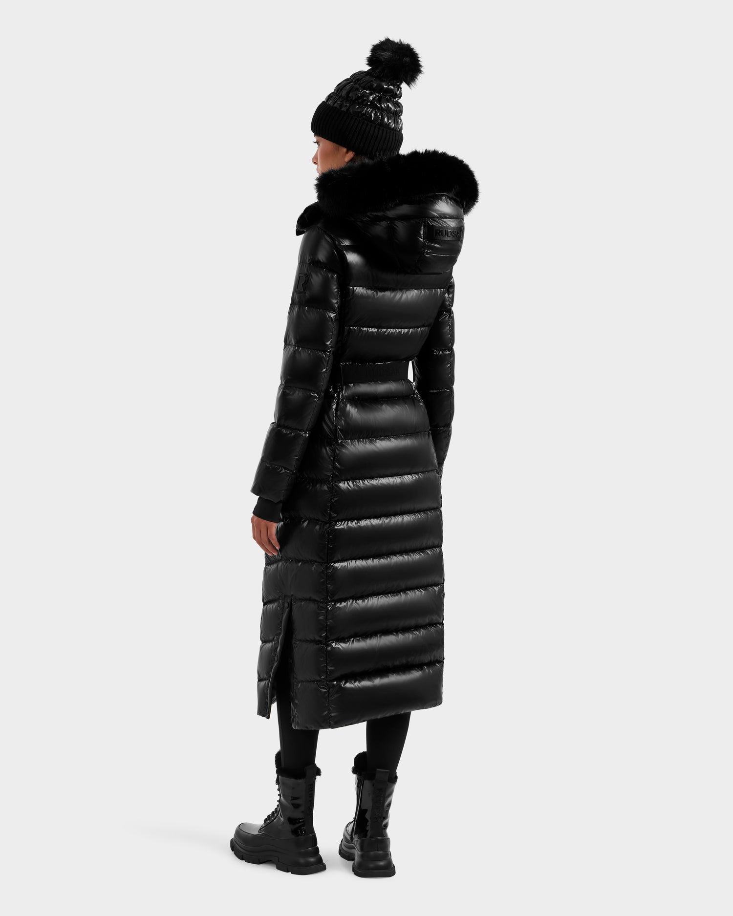 A person wearing the Rudsak ELSIE S - L124536 Black maxi down puffer coat with a fur-trimmed hood, black pom-pom beanie, black pants, and boots stands with their back to the camera.