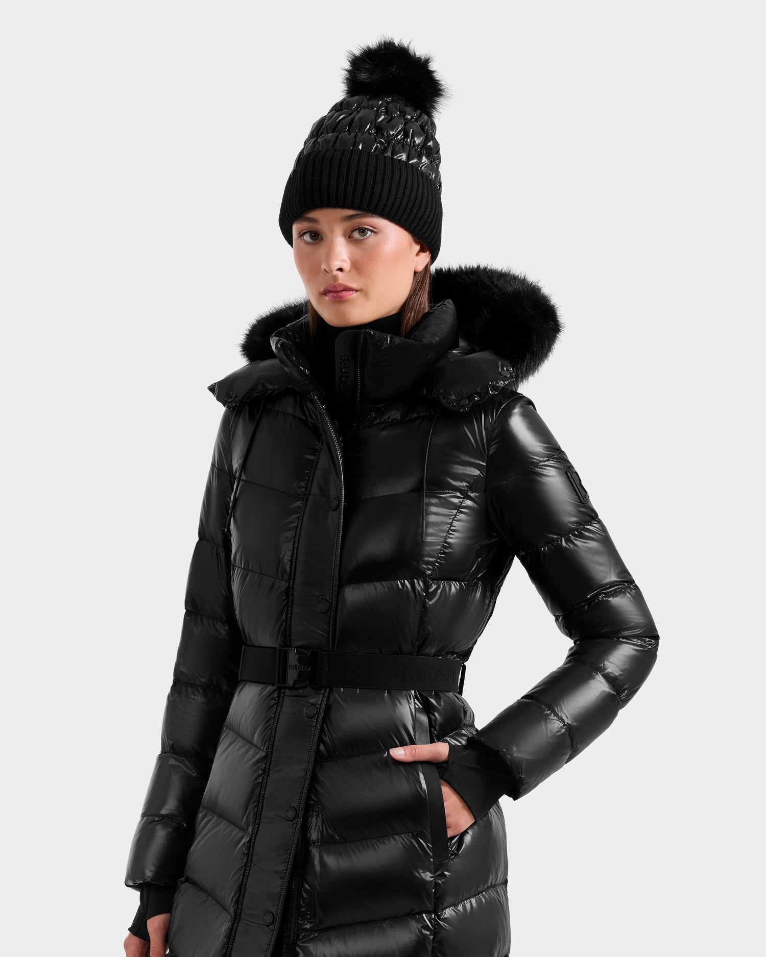A woman in the Rudsak ELSIE S - L124536 Black maxi down puffer with a fur-trimmed hood and a matching black knit pom-pom beanie stands against a plain light gray background.