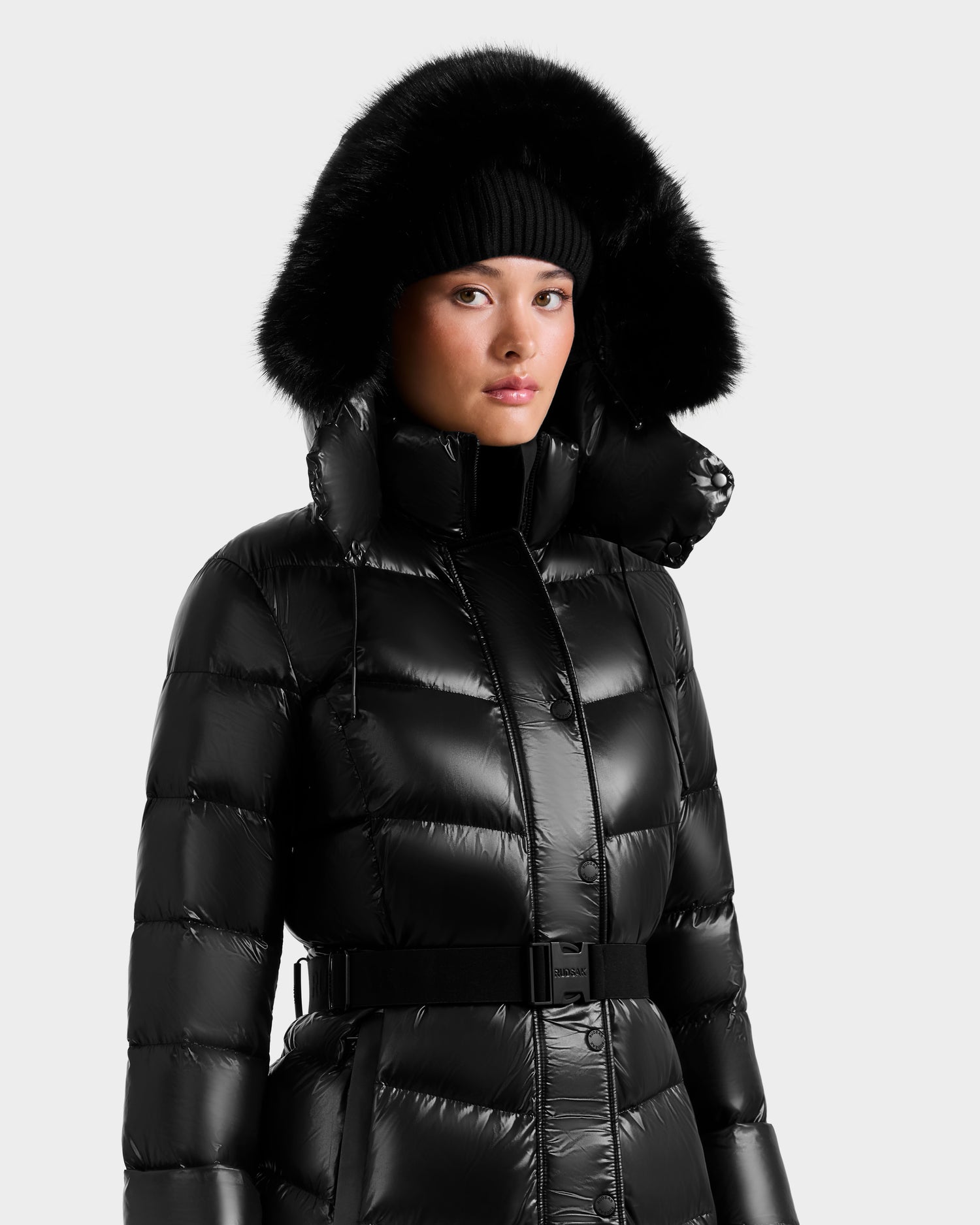 A person in the Rudsak ELSIE S - L124536 Black women’s maxi down puffer with a fur-trimmed hood and a black knit beanie, looking forward against a plain light background.