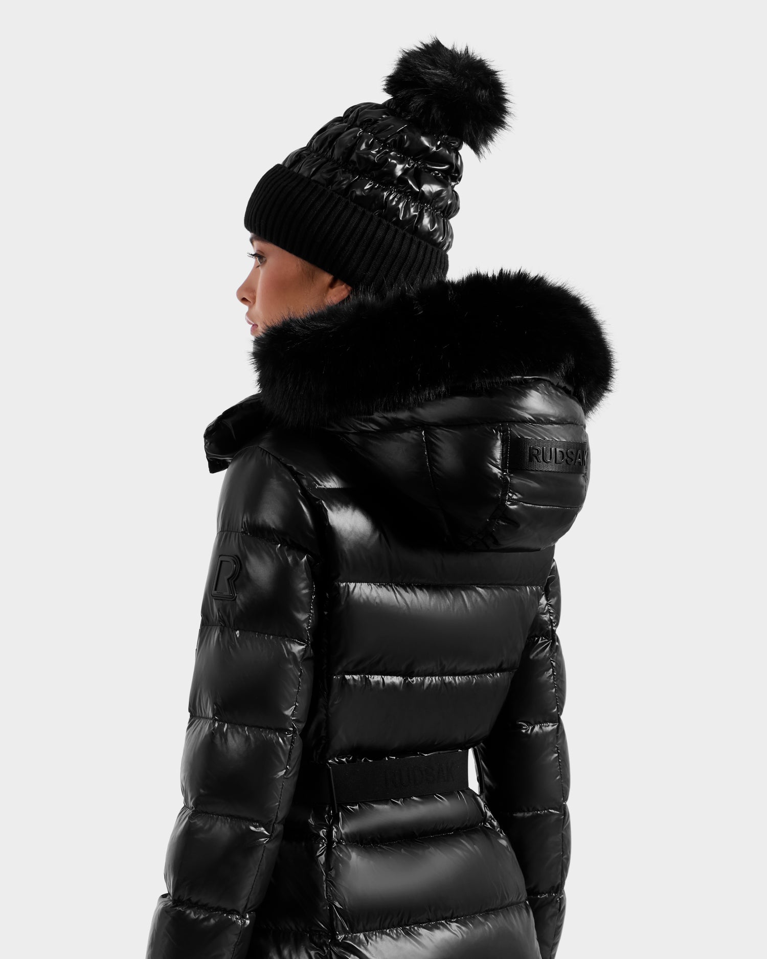 A person wearing the Rudsak ELSIE S - L124536 Black quilted down puffer jacket with a fur-trimmed hood and a matching black knit pom-pom beanie, shown from the back and side against a plain background.