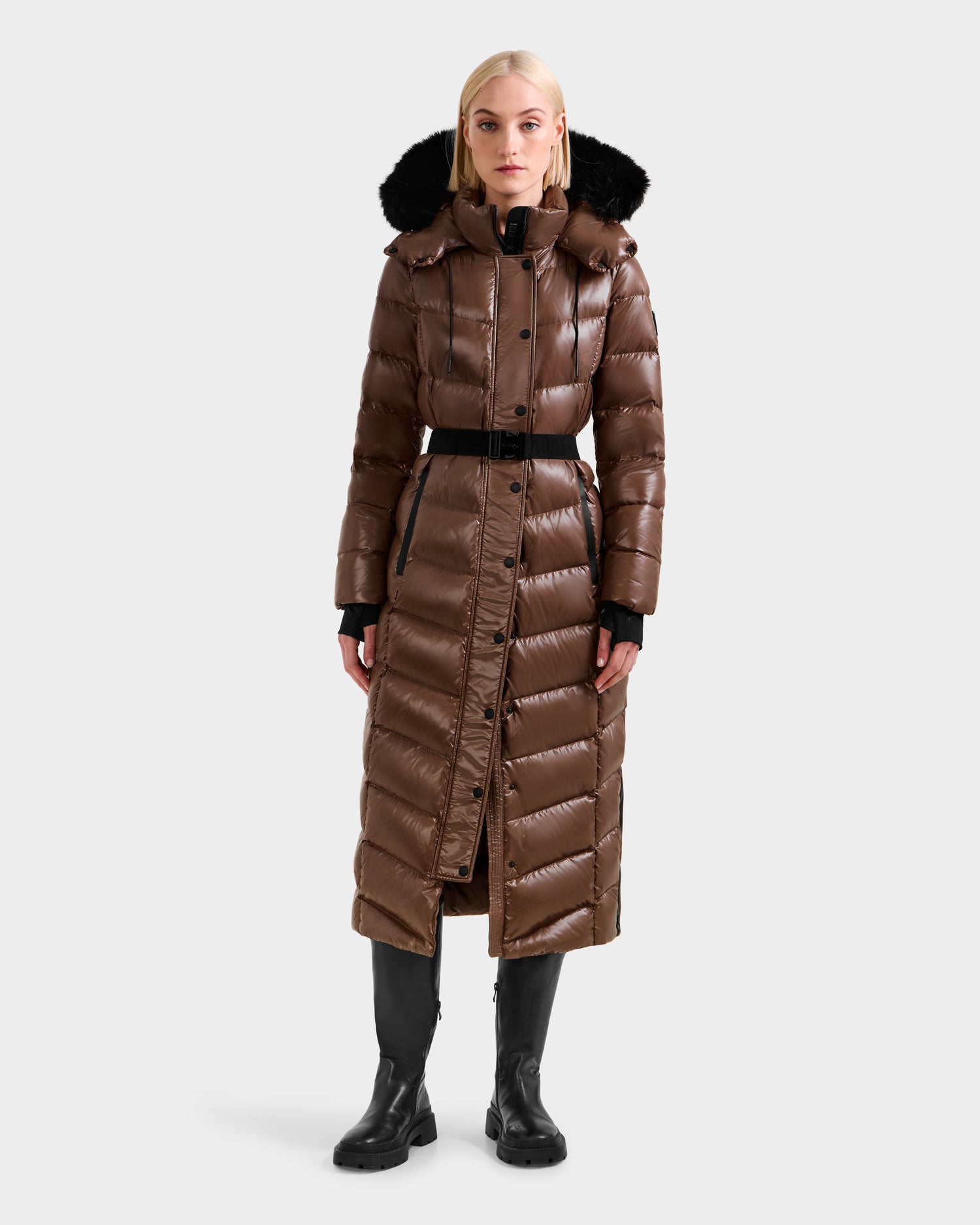 A woman stands facing forward against a plain light background wearing the Rudsak ELSIE S - L124536 Cacao women’s maxi down puffer coat with a black belt, black hood trim, and black boots.