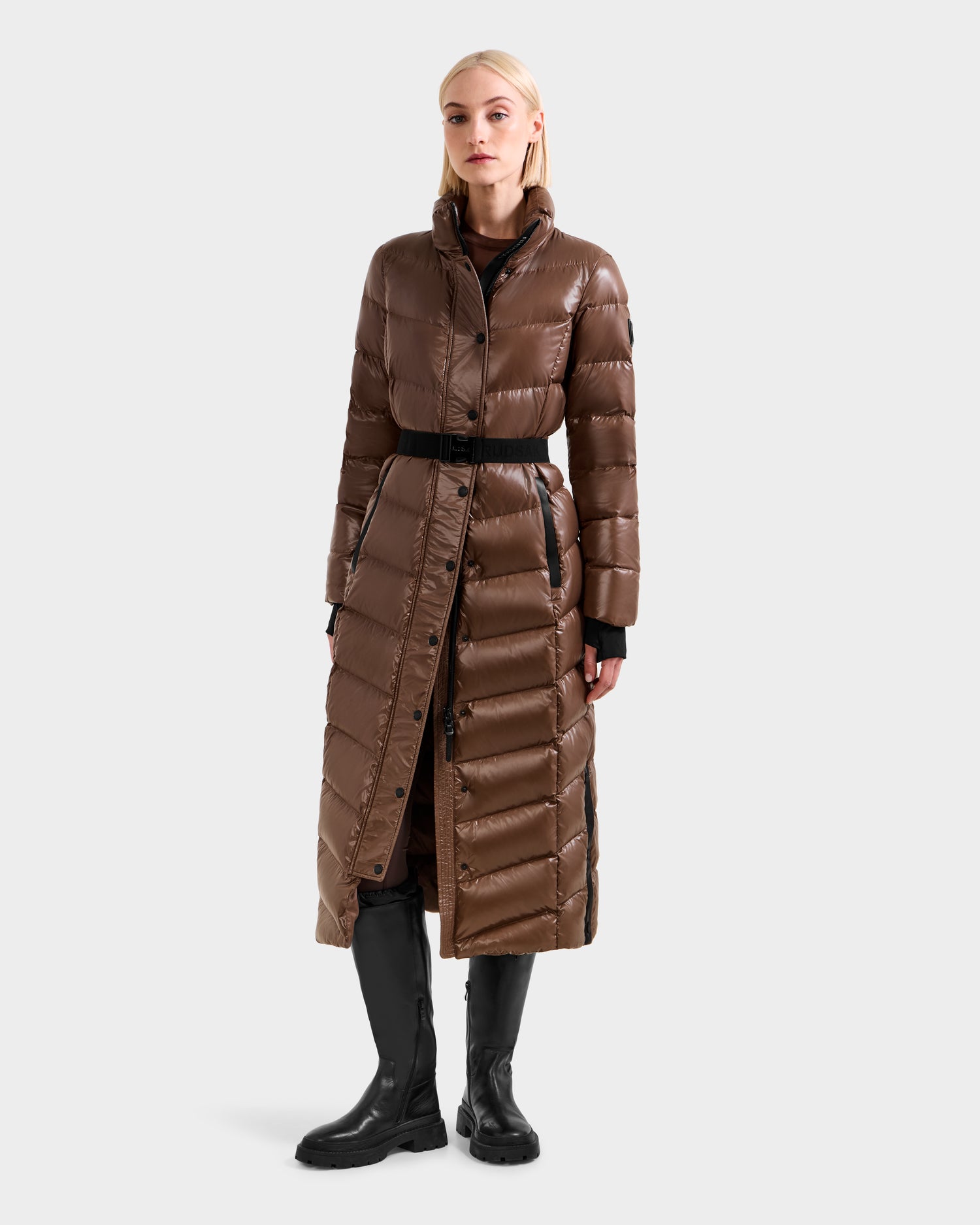A woman in the Rudsak ELSIE S - L124536 Cacao long belted maxi down puffer stands facing forward, styled over black clothing and knee-high black boots, against a plain light background.