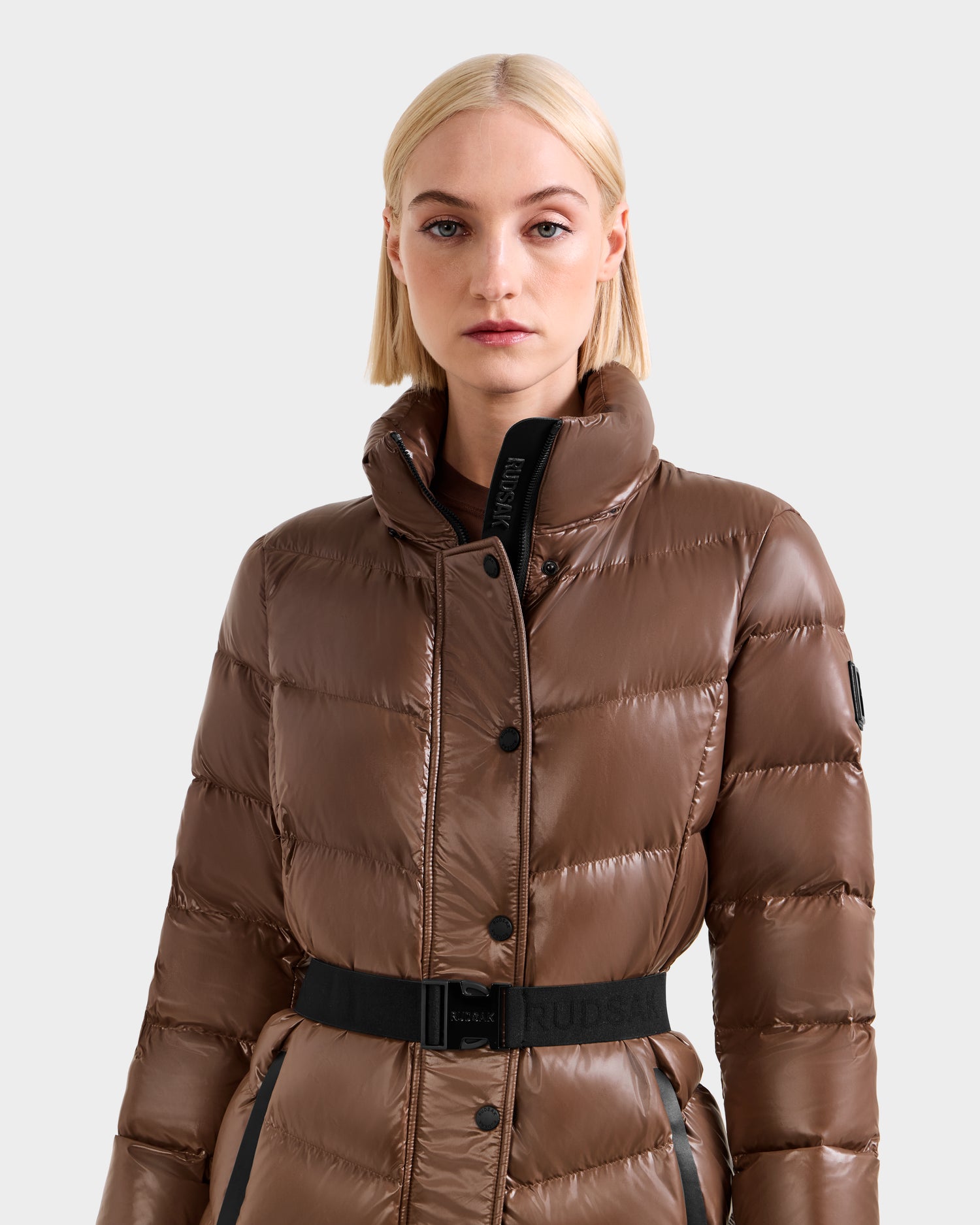 A woman with straight, blonde hair wears the shiny Rudsak ELSIE S - L124536 Cacao puffer jacket with a high collar and black belt, standing against a plain light background.