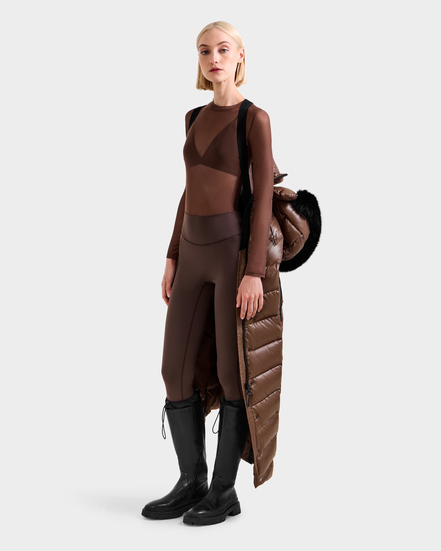 A woman in a sheer brown bodysuit and black knee-high boots stands holding the Rudsak ELSIE S - L124536 Cacao, an RDS-certified down winter coat with black fur trim, draped over her arm against a plain light background.