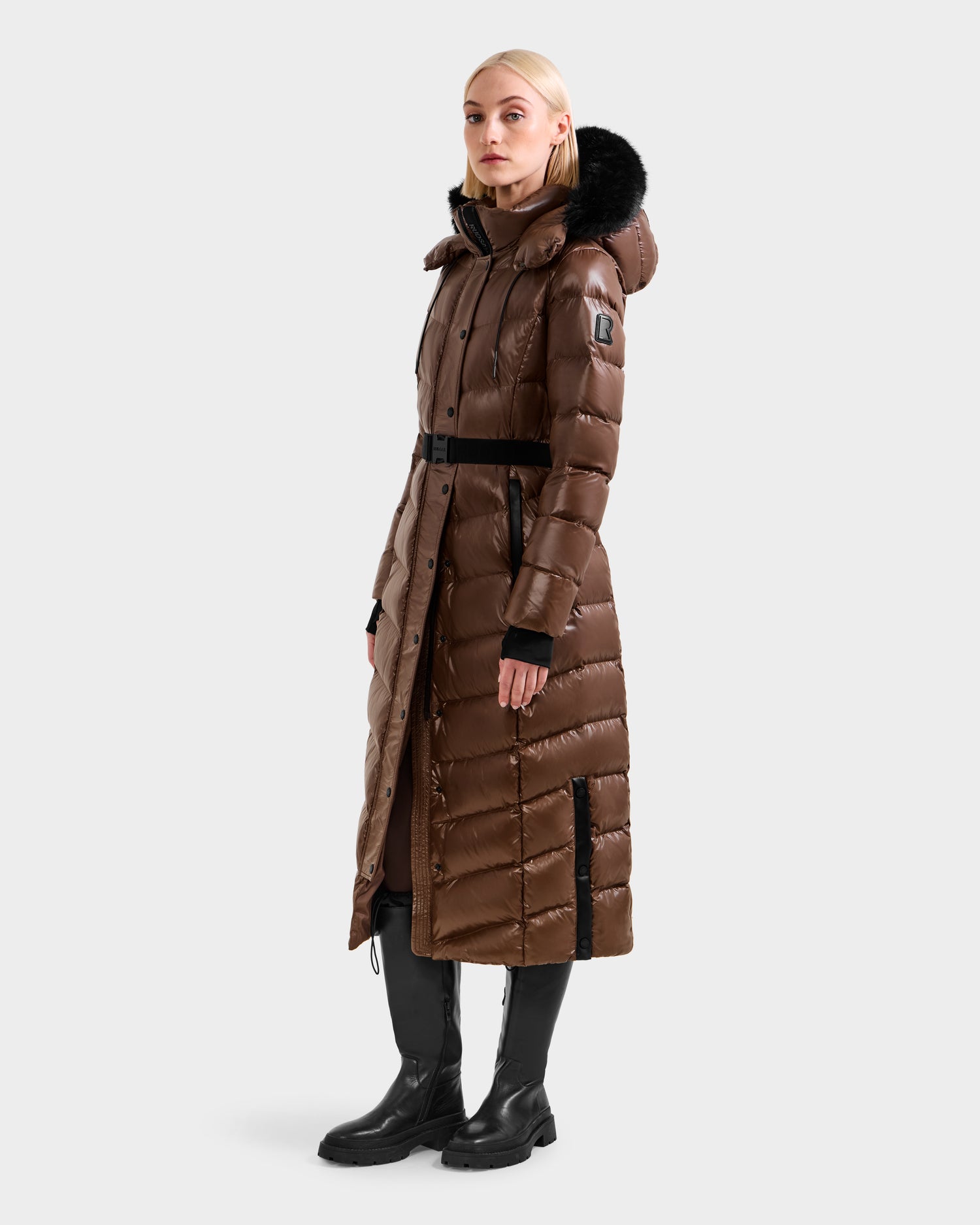 A woman wearing the Rudsak ELSIE S - L124536 Cacao maxi down puffer with a black fur-trimmed hood, black belt, and black boots stands against a plain light gray background.