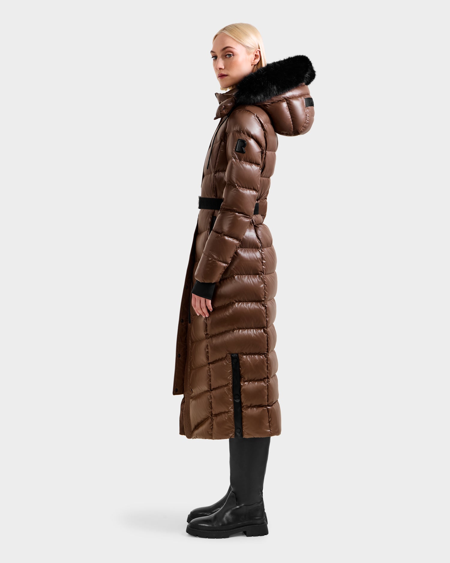 A blonde woman models the Rudsak ELSIE S - L124536 Cacao maxi down puffer coat with a black fur-trimmed hood, belted waist, and side vents, paired with black boots, standing in profile against a plain background.