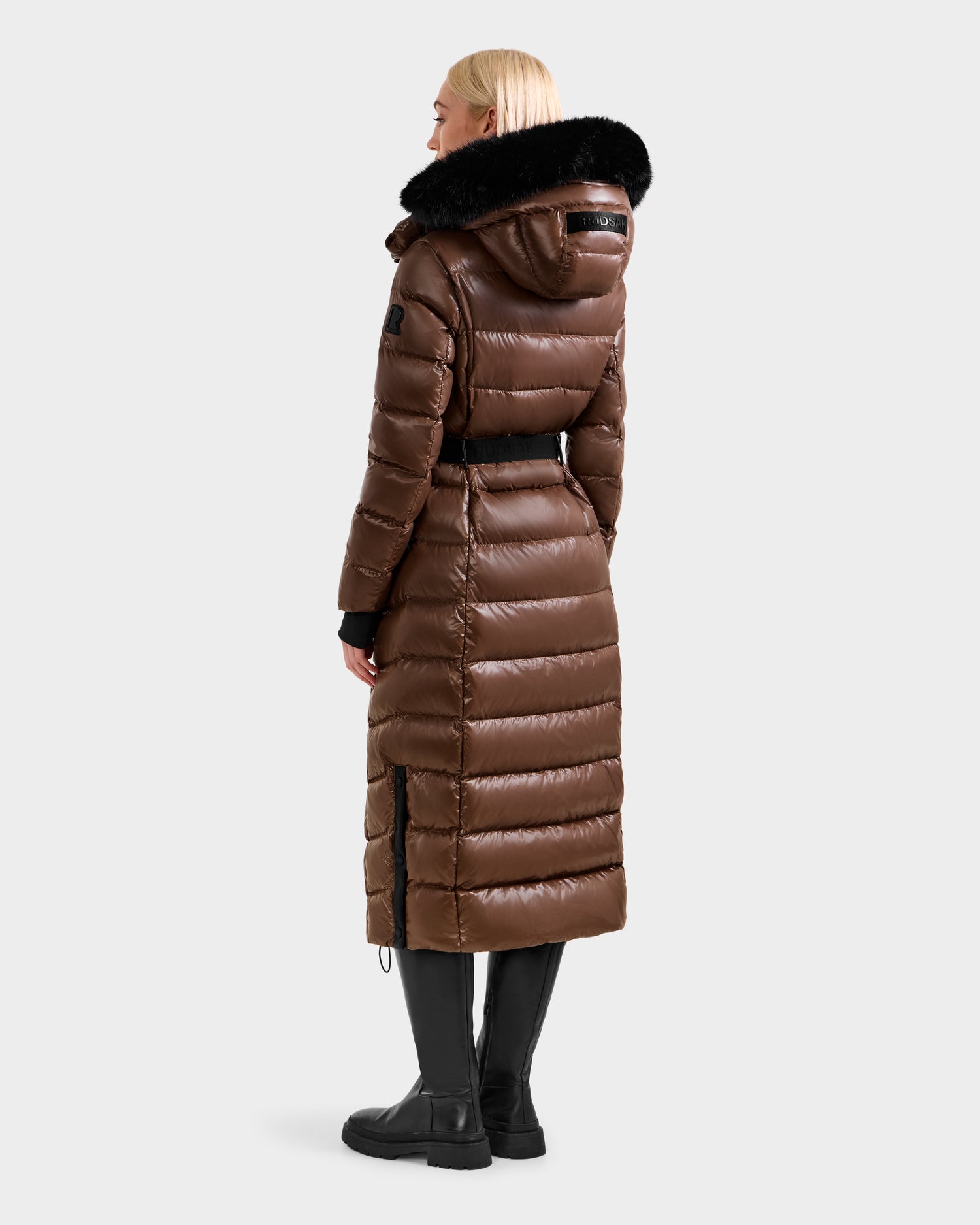 A blonde woman wears the Rudsak ELSIE S - L124536 Cacao, a shiny brown maxi down puffer with a black fur-trimmed hood, plus black leggings and boots, standing with her back to the camera against a plain background.
