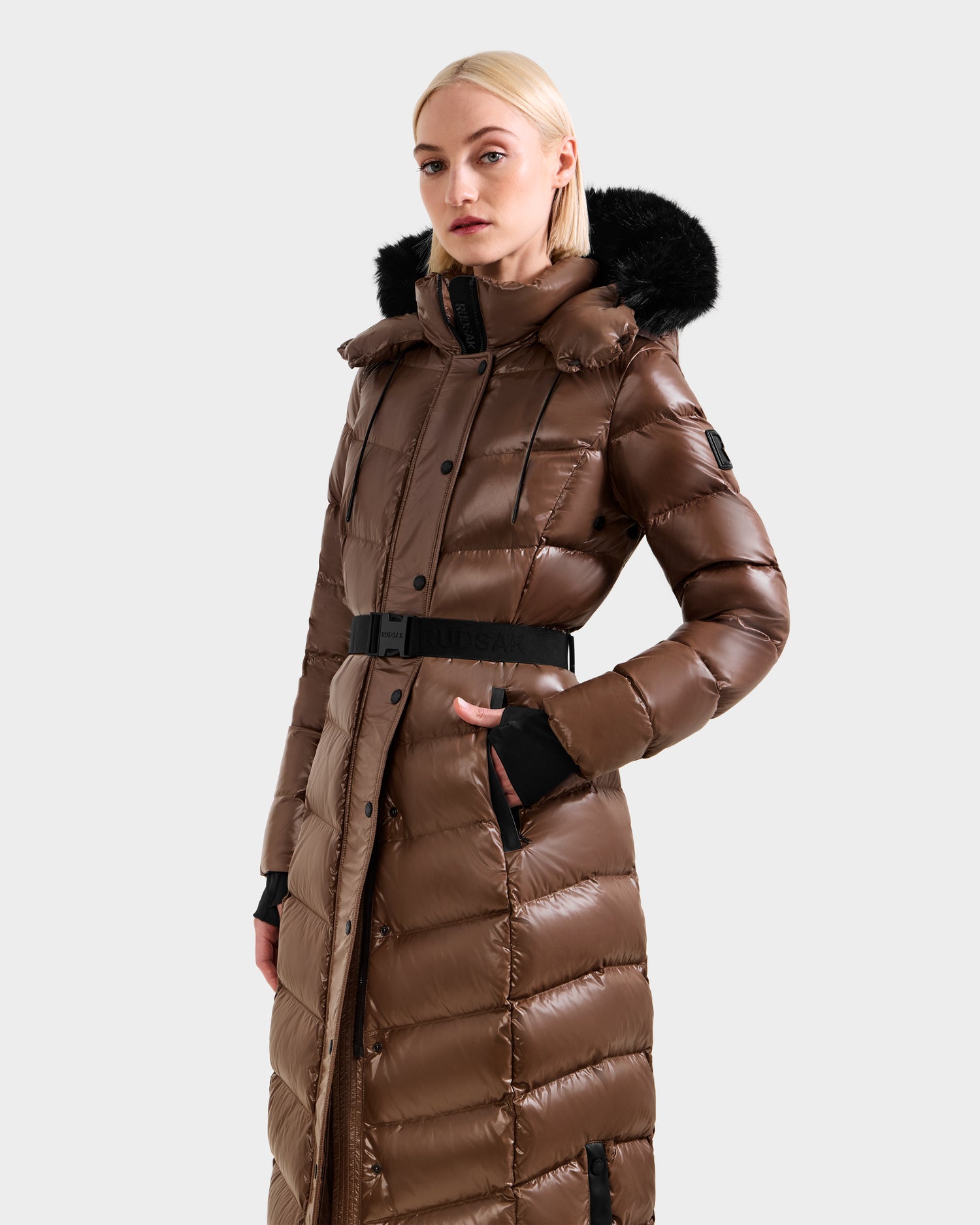 A woman wearing the Rudsak ELSIE S - L124536 Cacao maxi down puffer with a shiny brown finish, black fur-trimmed hood, and belted waist stands against a plain light gray background.