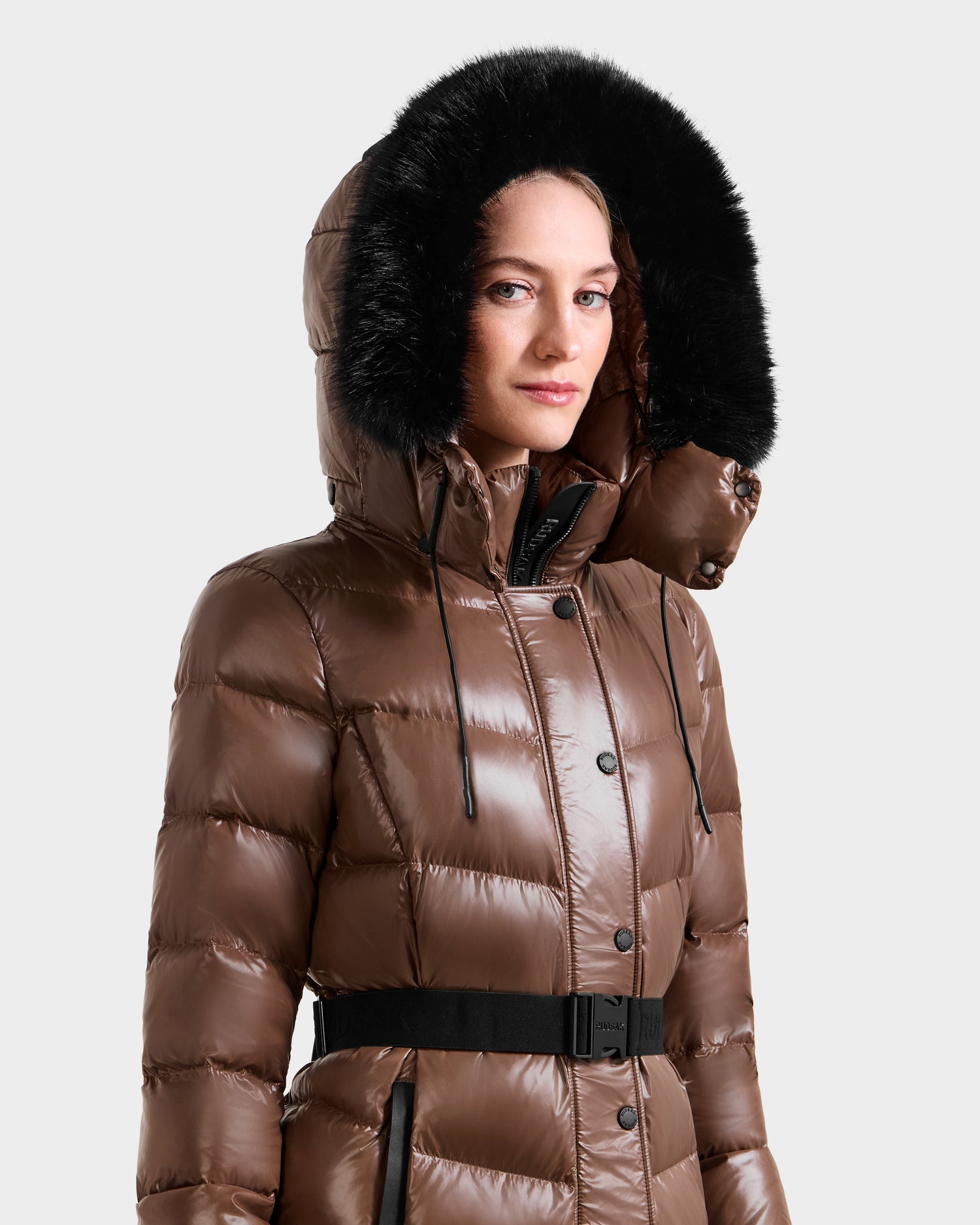 A woman models the Rudsak ELSIE S - L124536 Cacao, a shiny brown quilted down puffer coat with a black belted waist and large black faux fur-trimmed hood, standing against a plain light background.