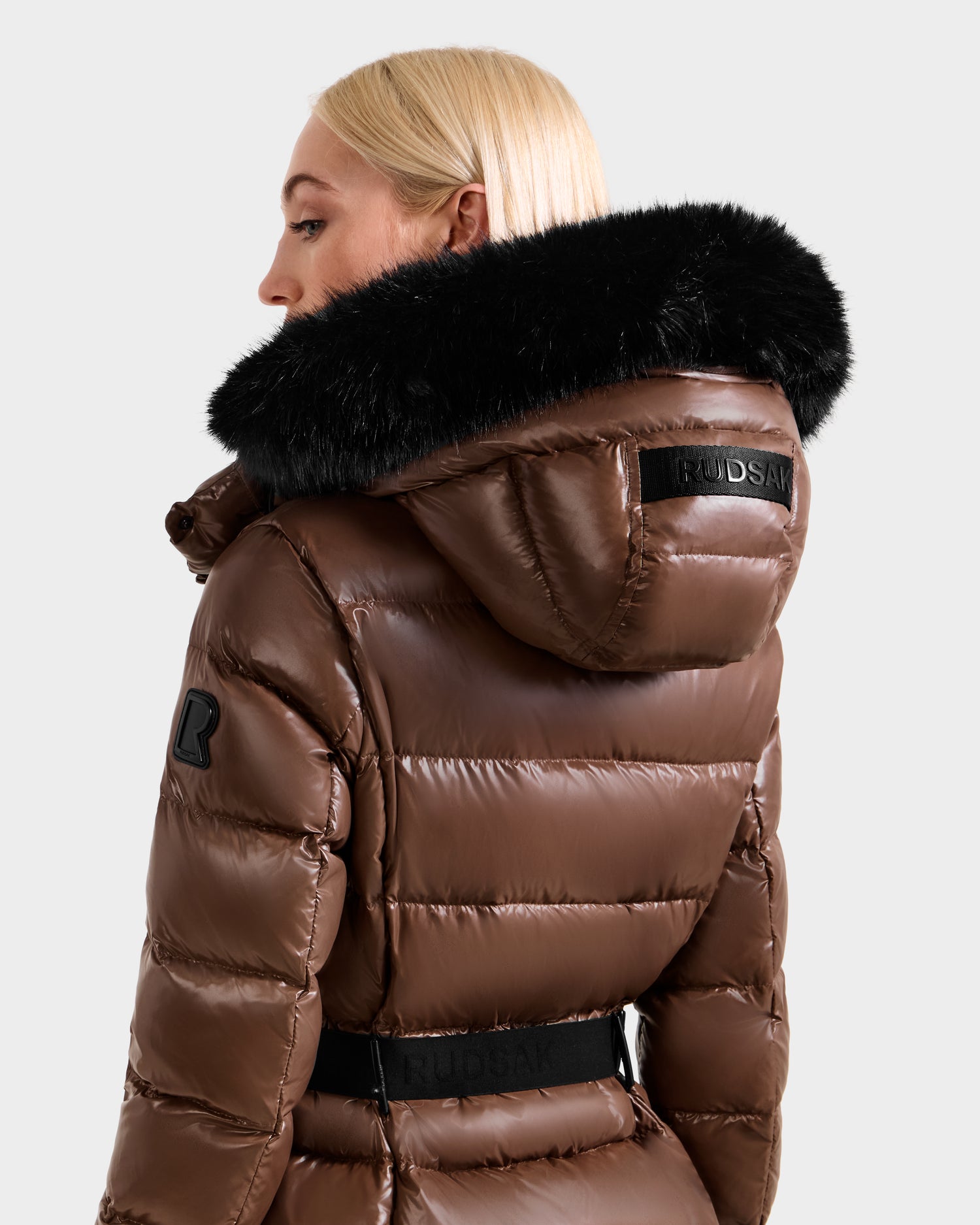 A woman with blonde hair is seen from the side and back, wearing the Rudsak ELSIE S - L124536 Cacao maxi down puffer coat with a large black fur-trimmed hood. The coat features patches and visible Rudsak branding on the hood and belt.