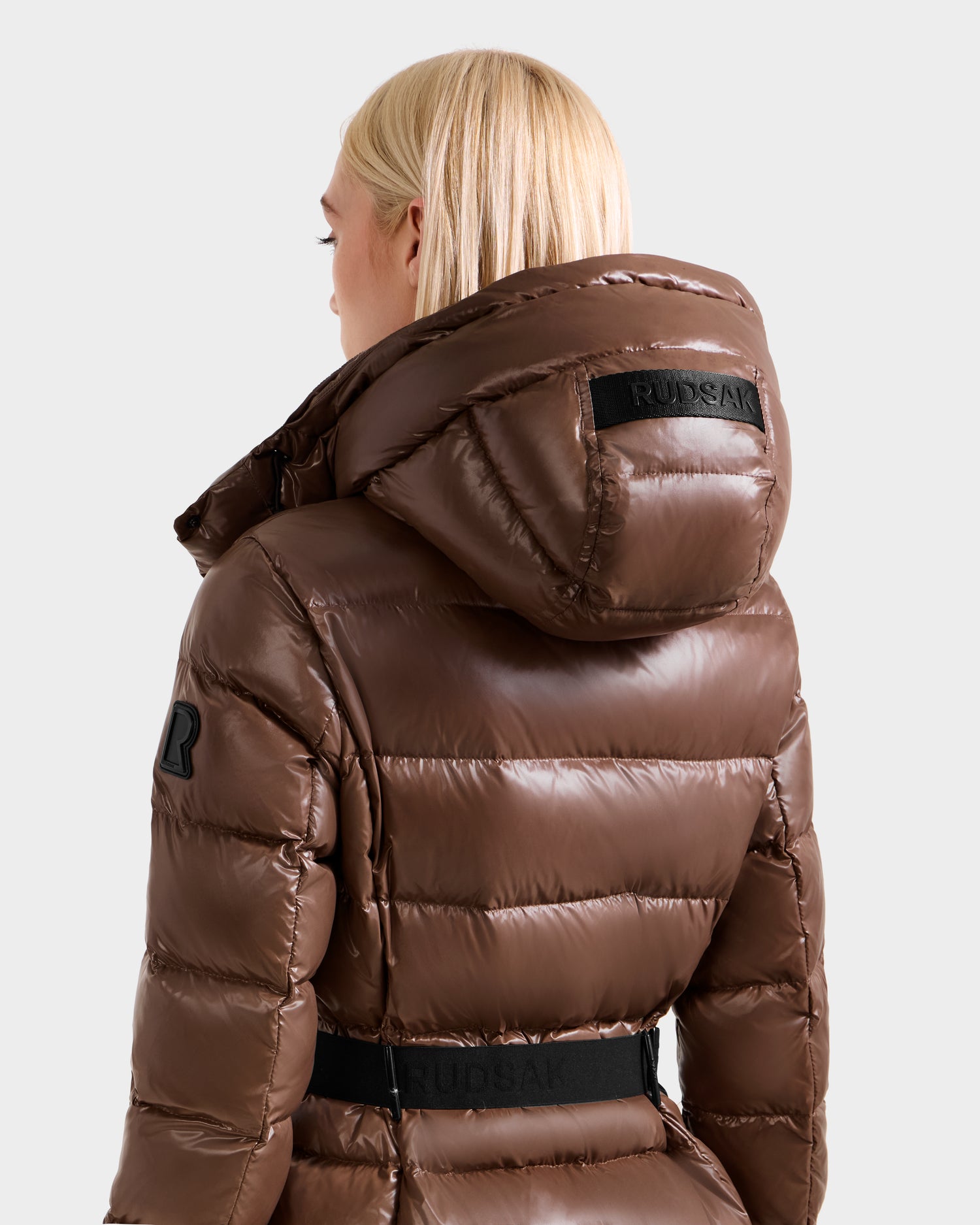 A woman with straight blonde hair models the Rudsak ELSIE S - L124536 Cacao maxi down puffer jacket with a hood and black RUDSAK labels, shown from the back and side against a plain background.