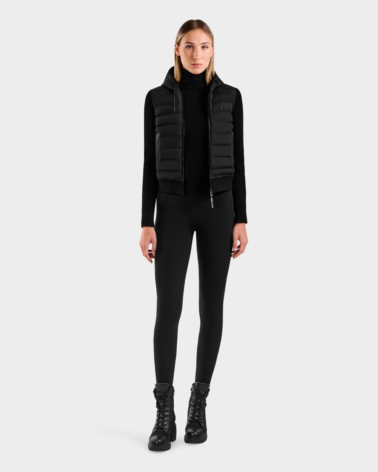 A woman faces forward wearing the EMILY - L125515 Black hooded down bomber by Rudsak over a black long-sleeve top, black leggings, and lace-up boots, set against a plain light gray background.
