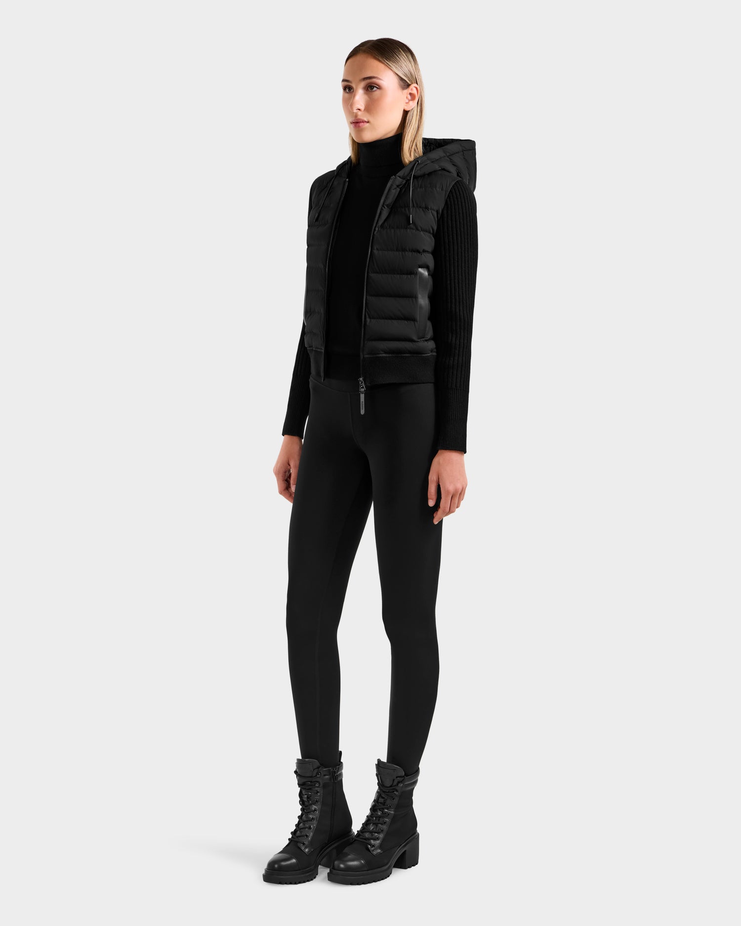 A woman stands facing forward against a plain light background, wearing the Rudsak EMILY - L125515 Black women’s hooded down bomber with a black long-sleeve top, leggings, and lace-up boots.