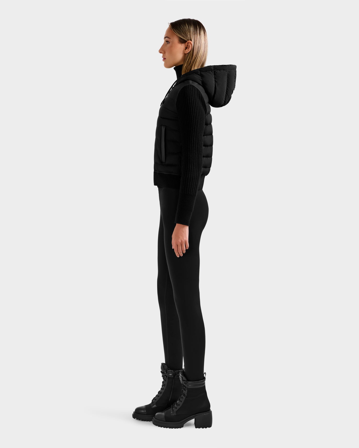 A woman in profile wears the Rudsak EMILY - L125515 Black hooded down bomber, styled with a black long-sleeve top, leggings, and lace-up boots, set against a plain light gray background.